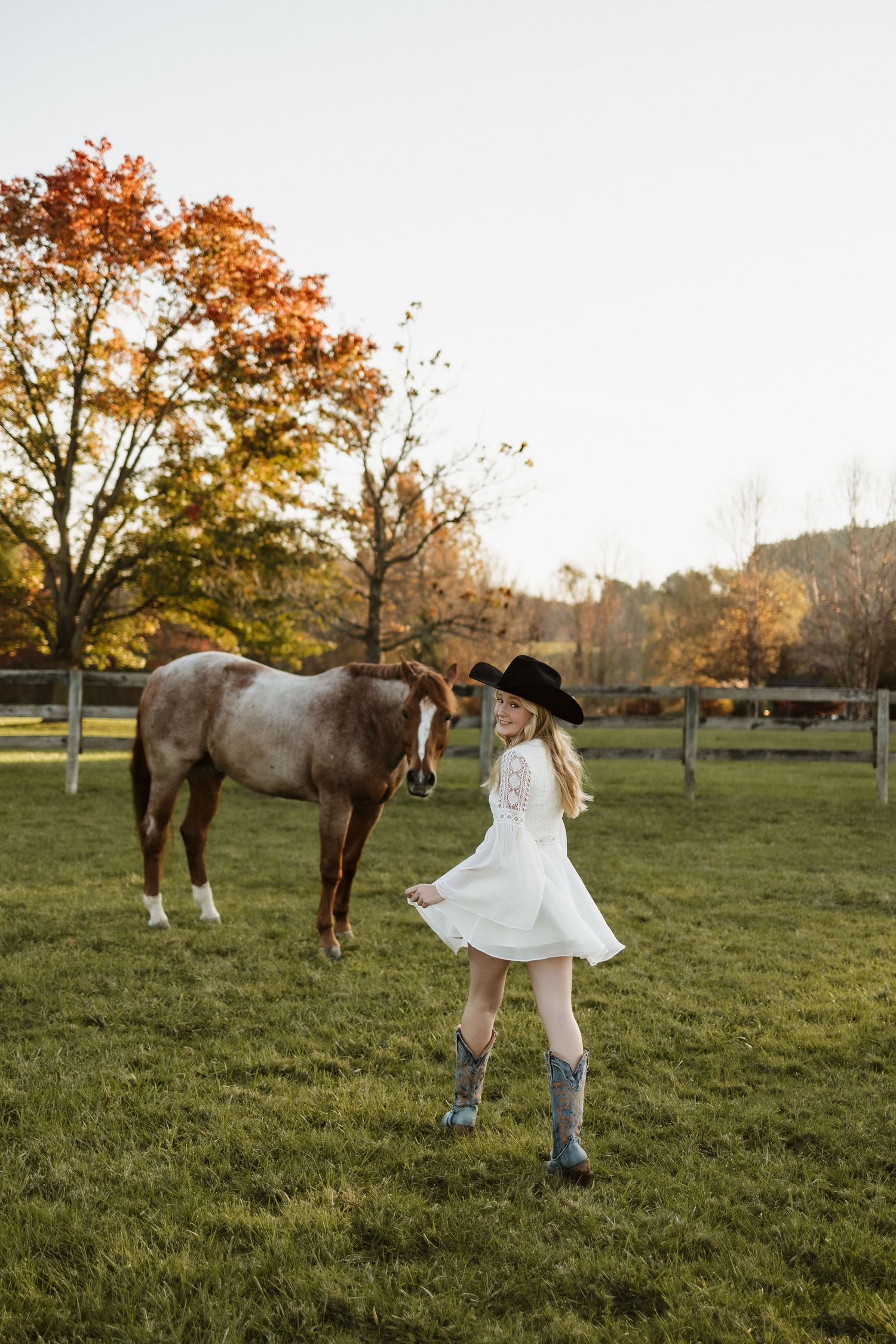 A person in a white dress and cowboy boots twirls in a field with a brown horse on an autumn day.