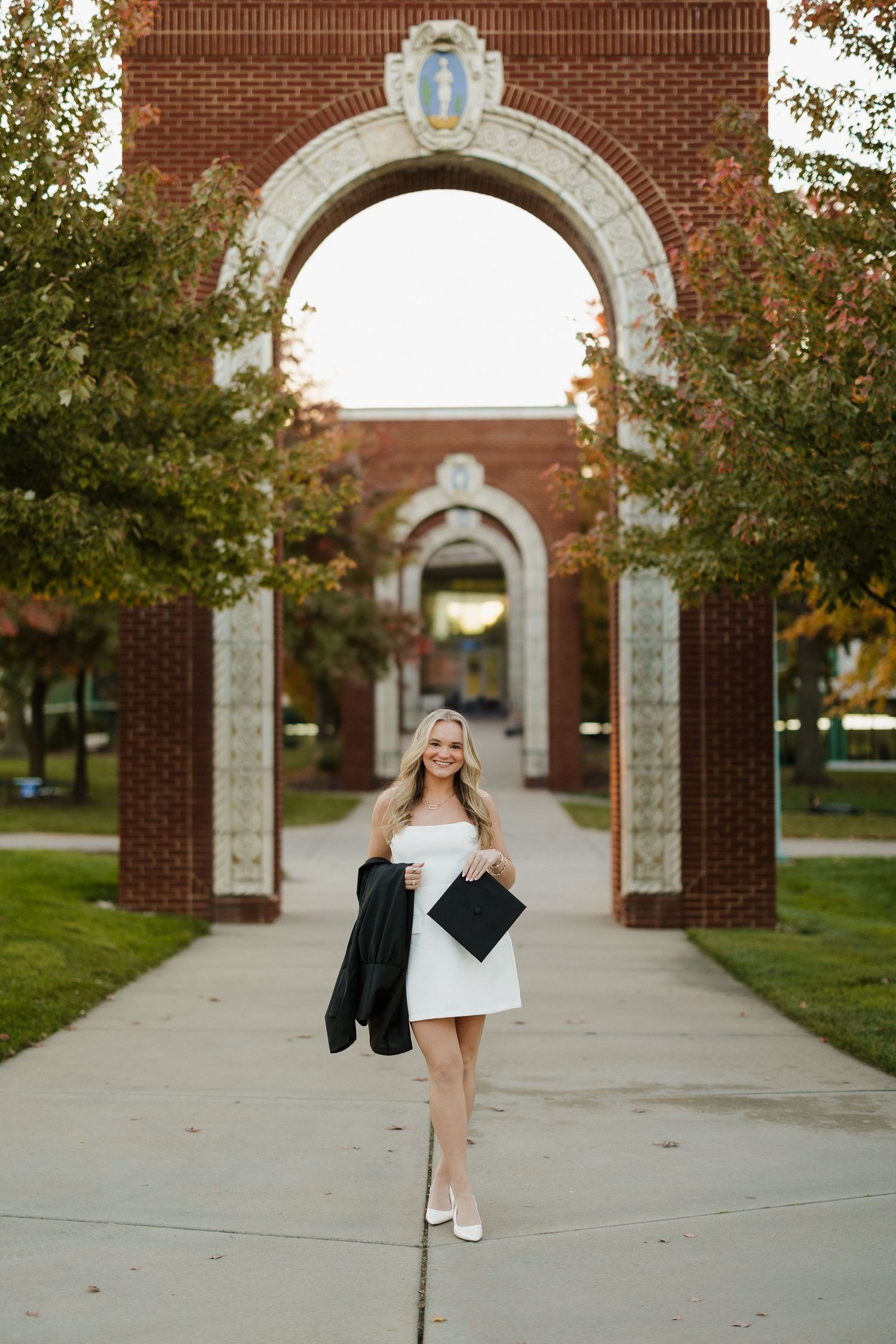 Graduate in white dress walks under ornate brick archway on college campus.