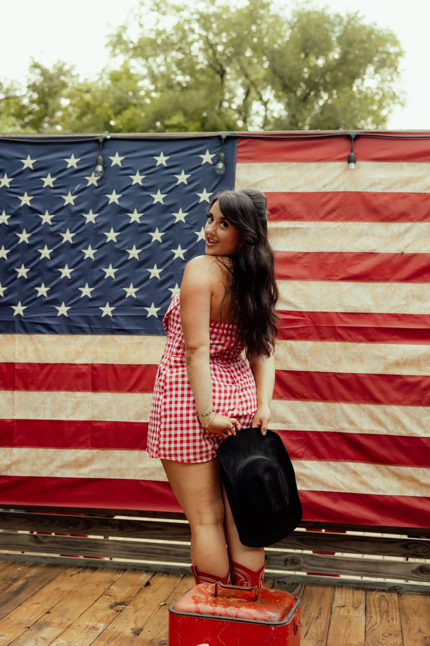 Woman in red gingham top poses in front of American flag on wooden deck in patriotic photoshoot series.