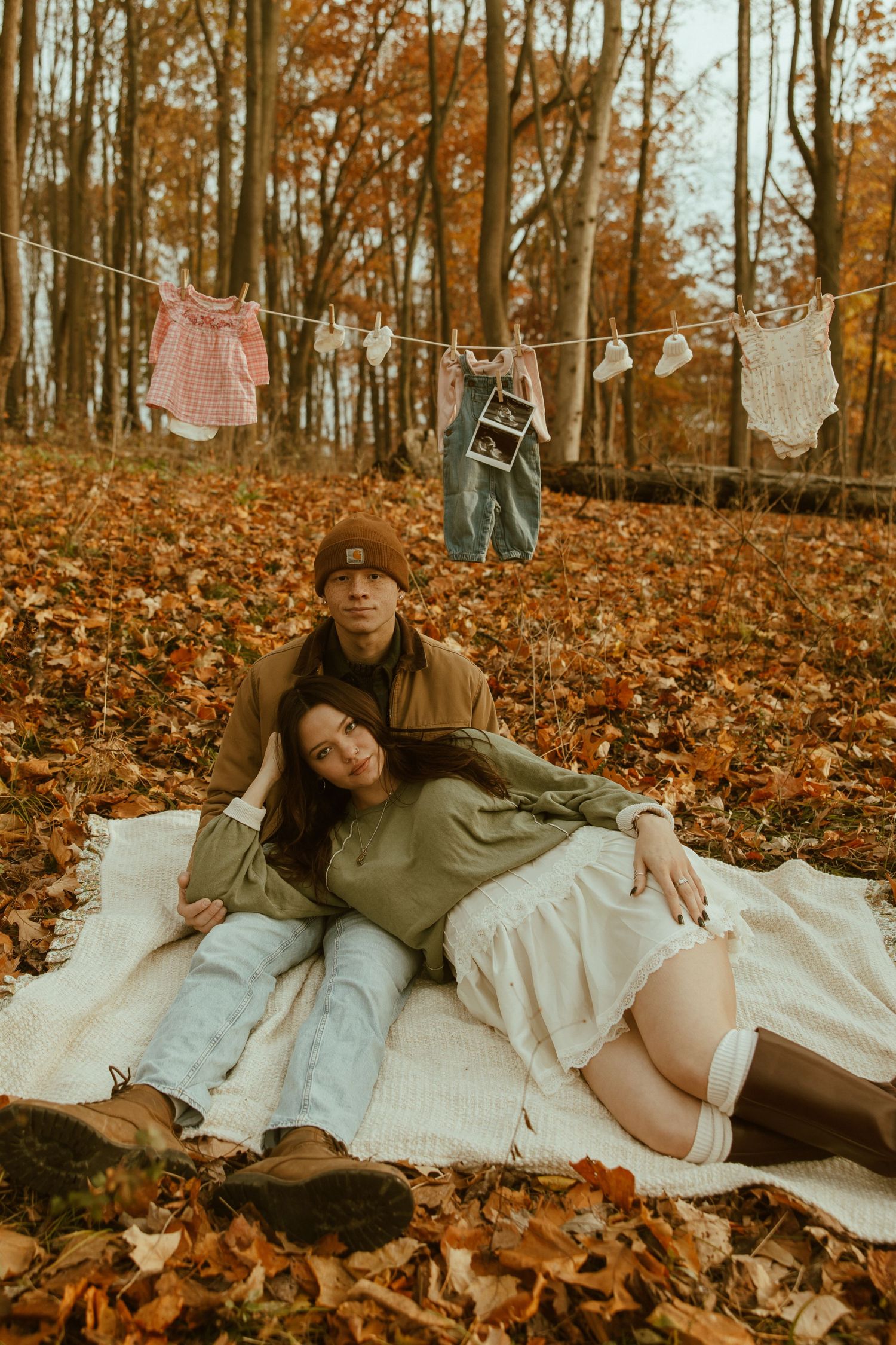 A romantic autumn photoshoot on a blanket surrounded by fallen orange leaves in a forest setting.