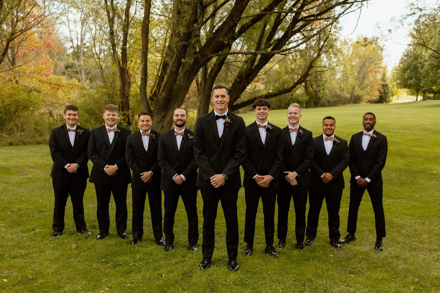 A wedding party in formal black attire poses outdoors in a line against an autumn landscape with trees.