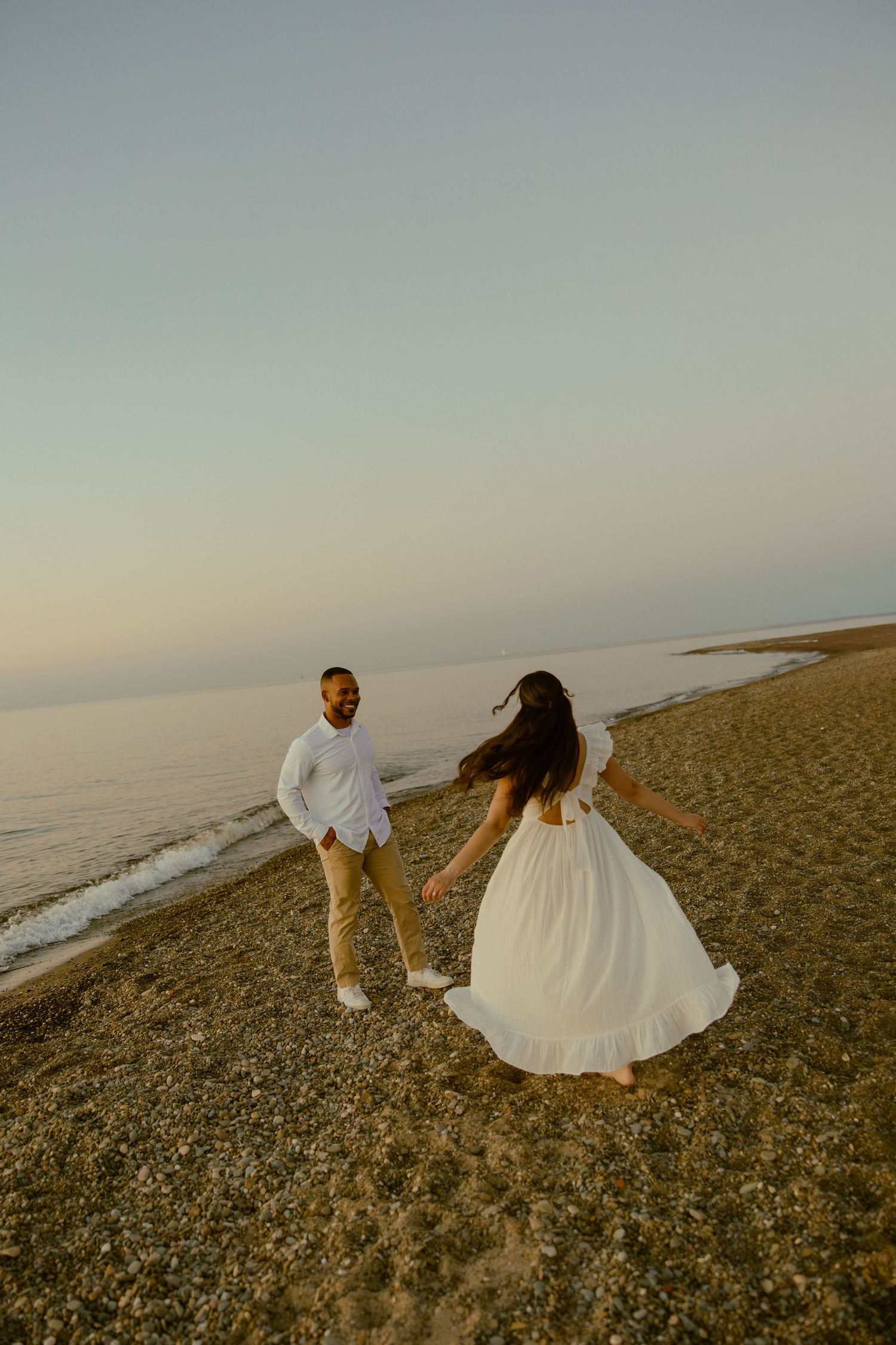 Couple in wedding attire walking along a scenic coastal beach at sunset with waves crashing in the background.