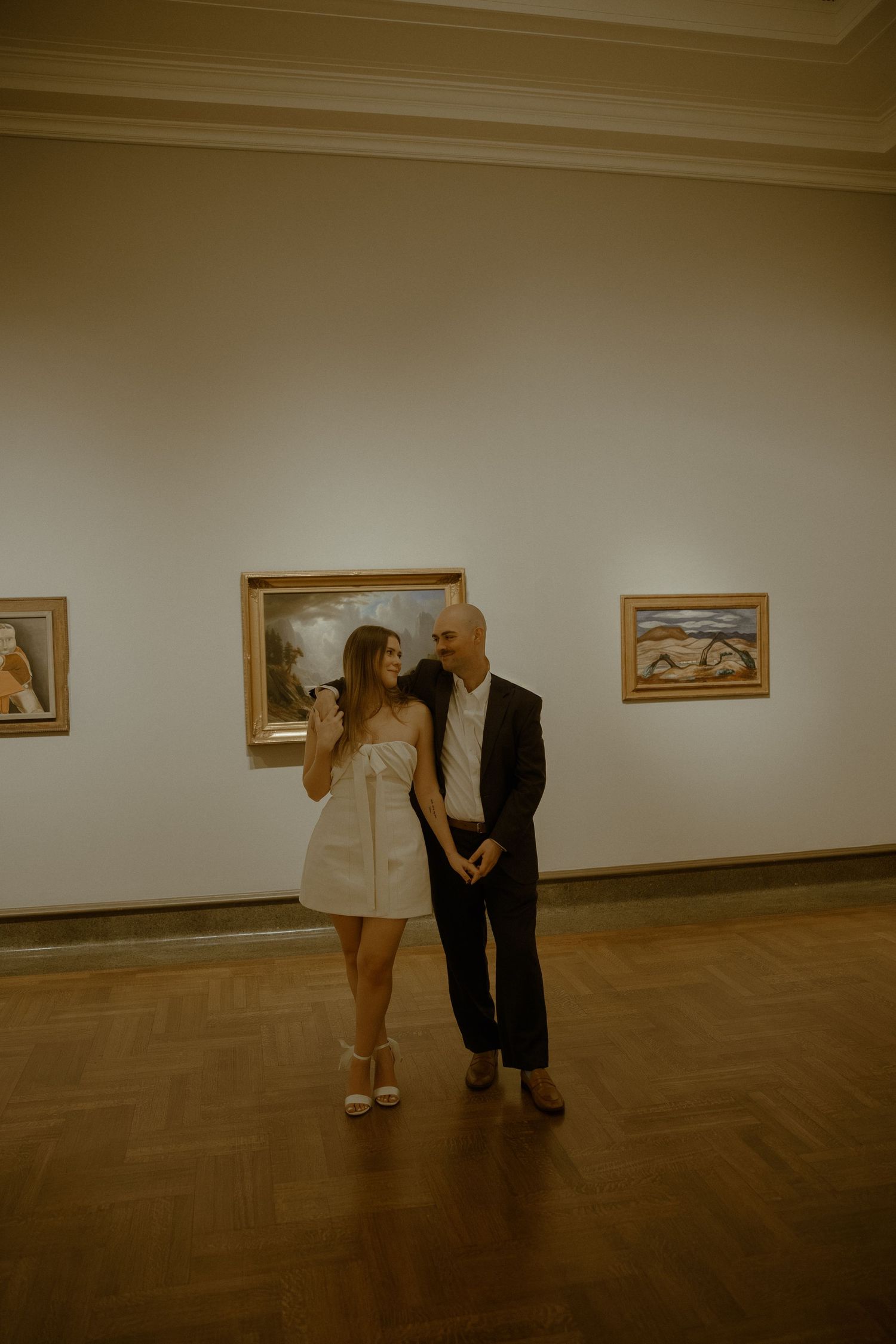 Two people stand together in an art gallery with framed paintings on white walls and a polished wooden floor.