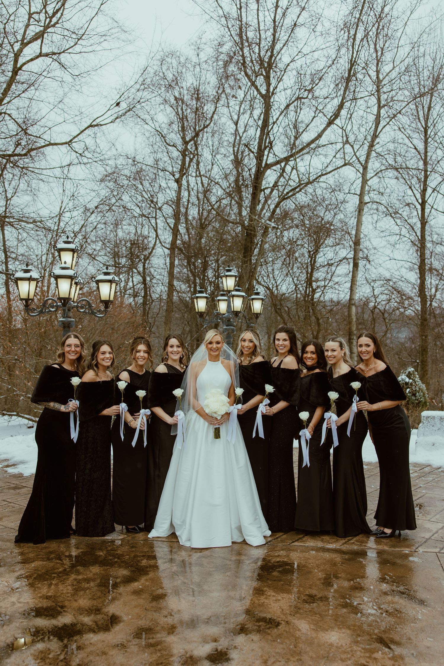 Wedding party in black dresses and white gown pose together in snowy winter setting with vintage lantern decorations.