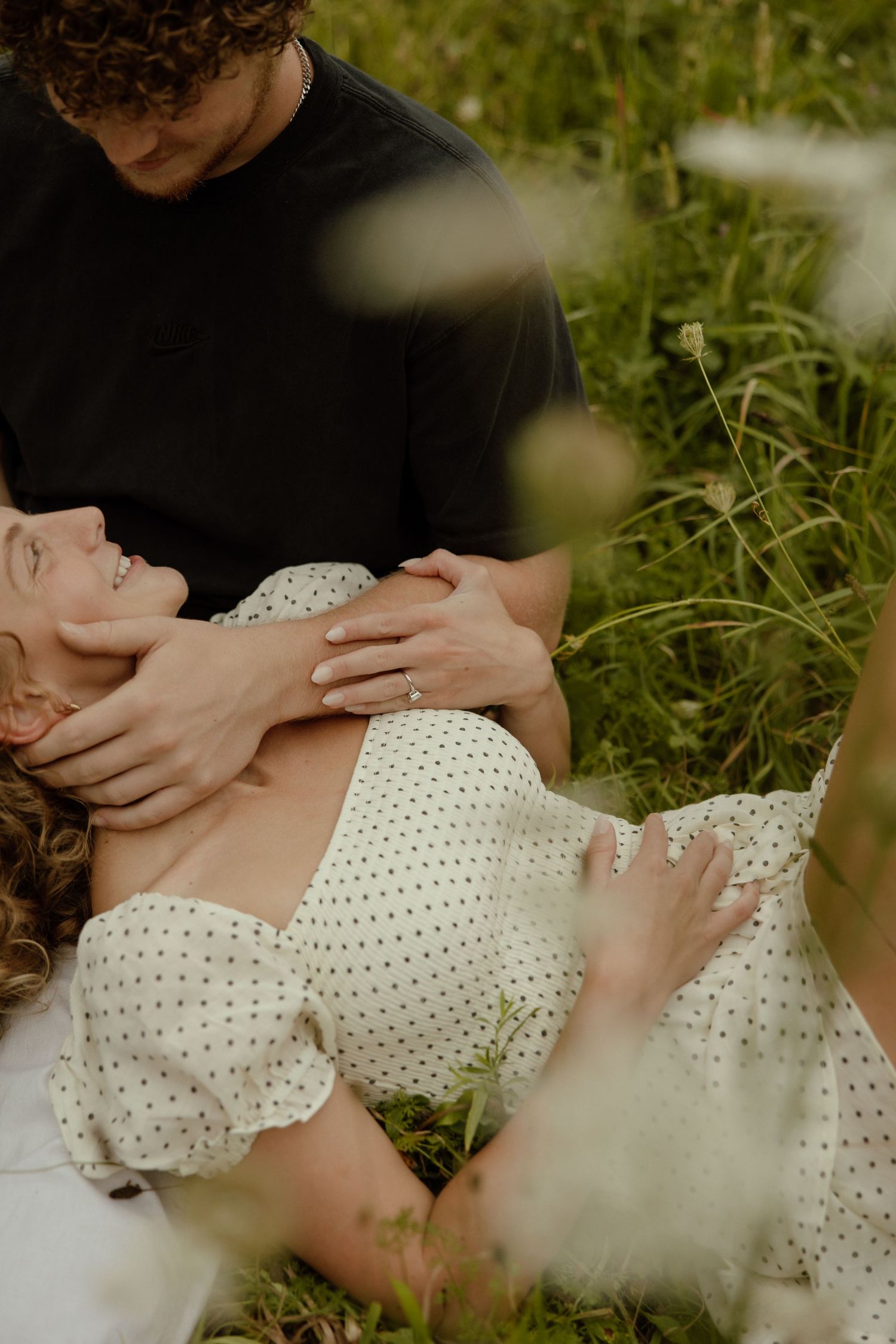 A romantic couple embraces on grass while wearing white polka dot clothing in an intimate outdoor setting.