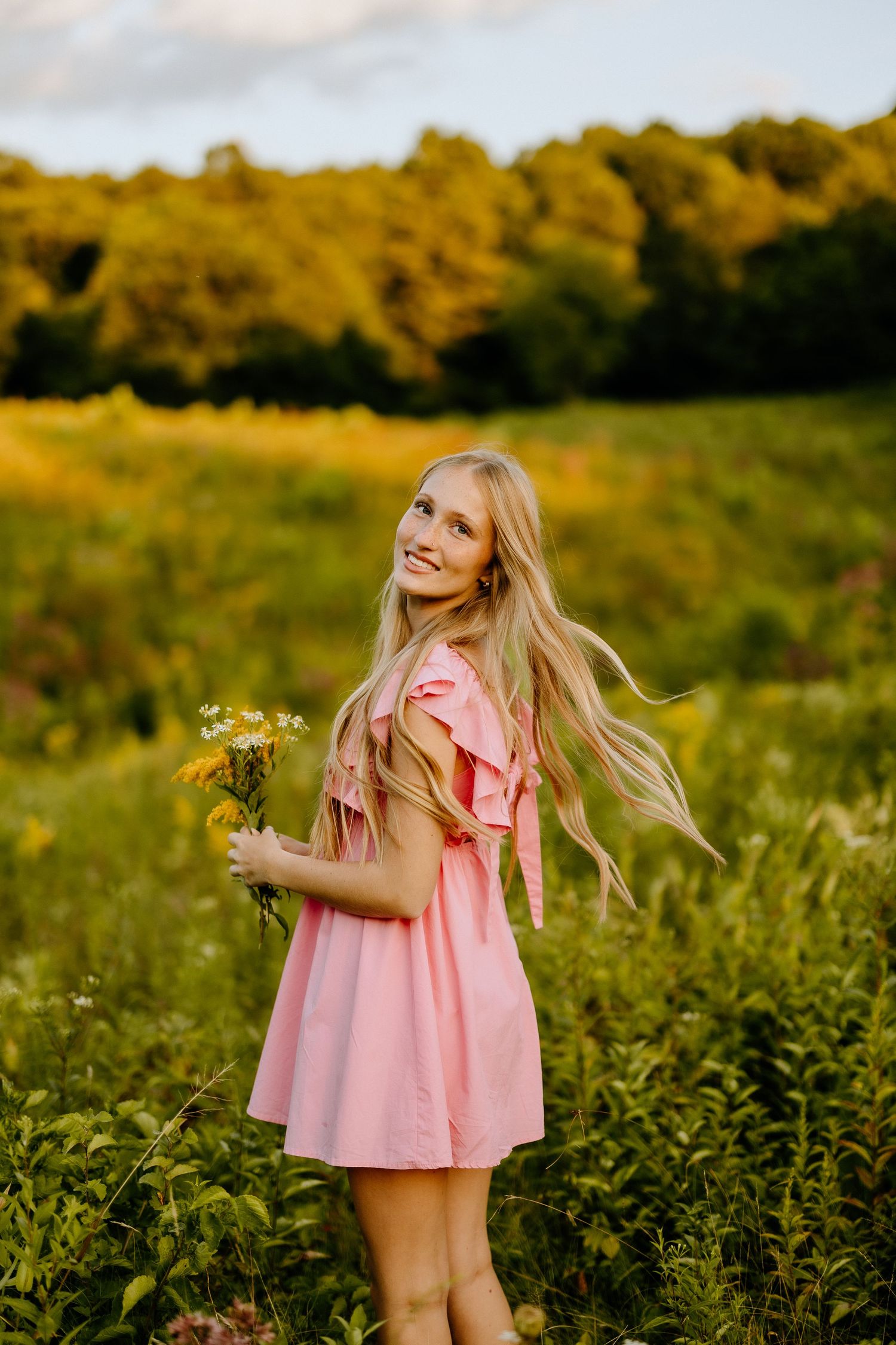 A pink sundress catches wind in a golden field at sunset while holding wildflowers.