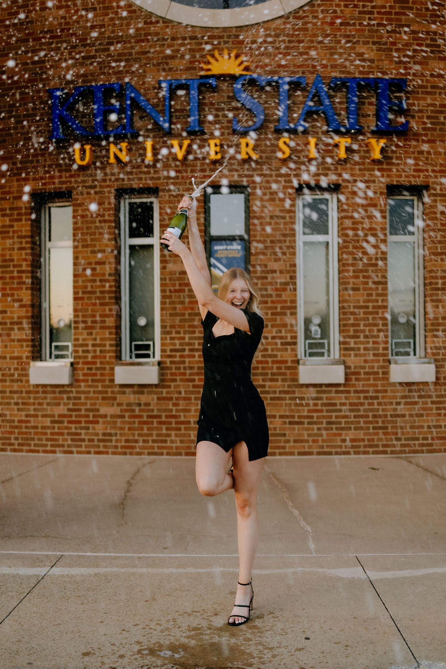 A graduate in a black dress jumps joyfully in front of Kent State University brick building.