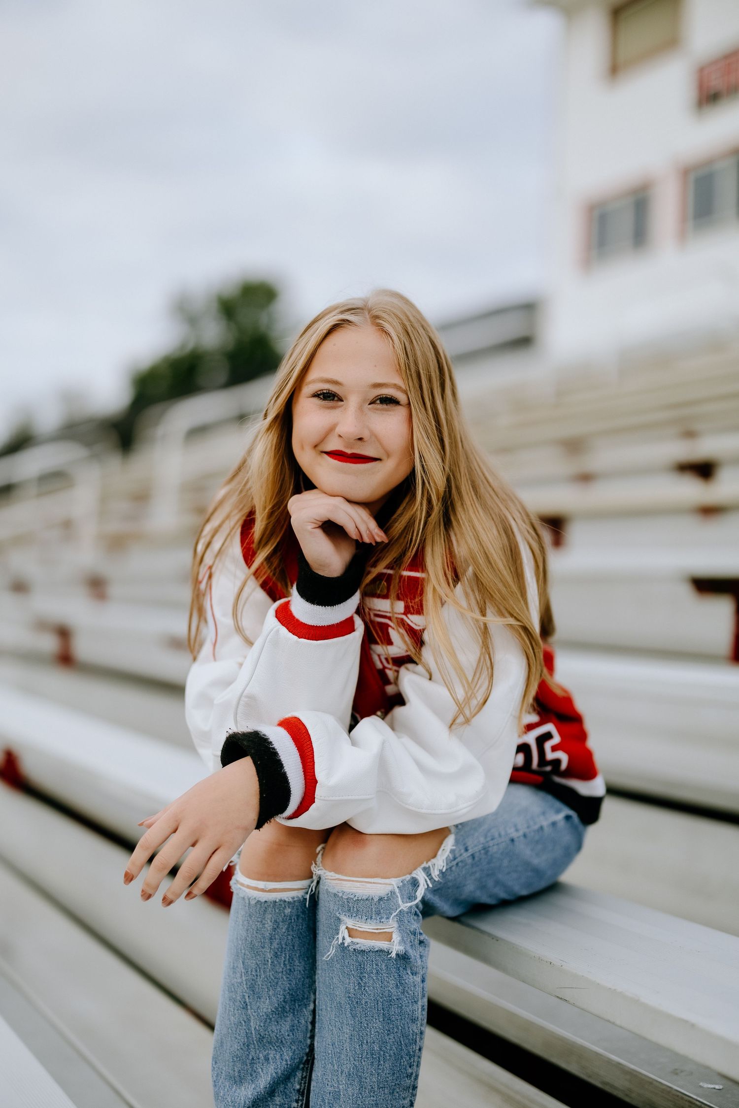 Person in varsity jacket and ripped jeans poses casually on bleachers.