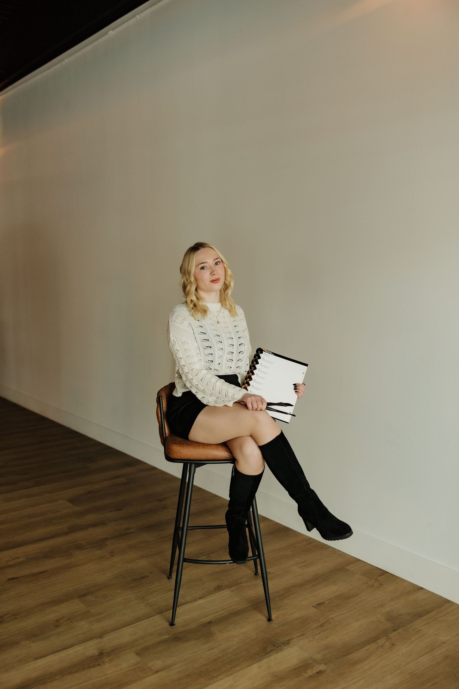 Stylish winter portrait in white sweater and knee-high boots holding notebook on wooden stool.