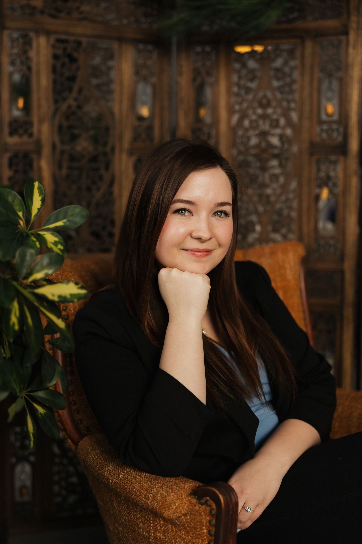 Portrait of person seated in brown chair against ornate wooden paneled wall with plants.