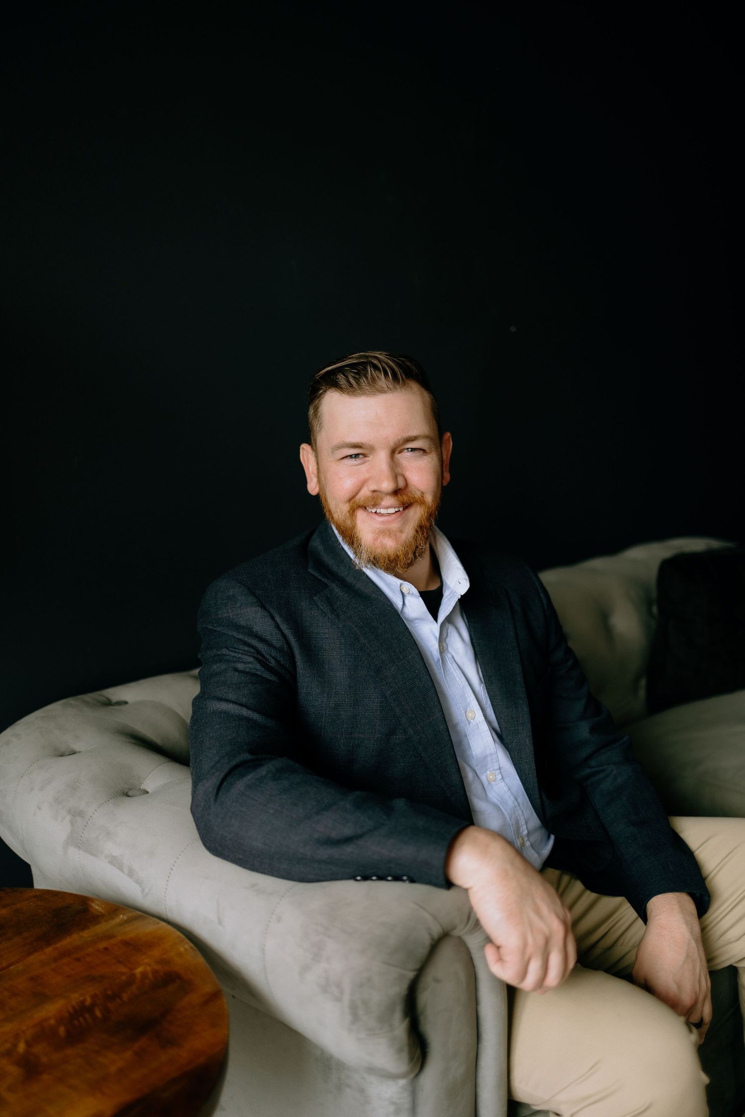 Professional in dark suit and white shirt sitting casually on gray armchair against dark backdrop.