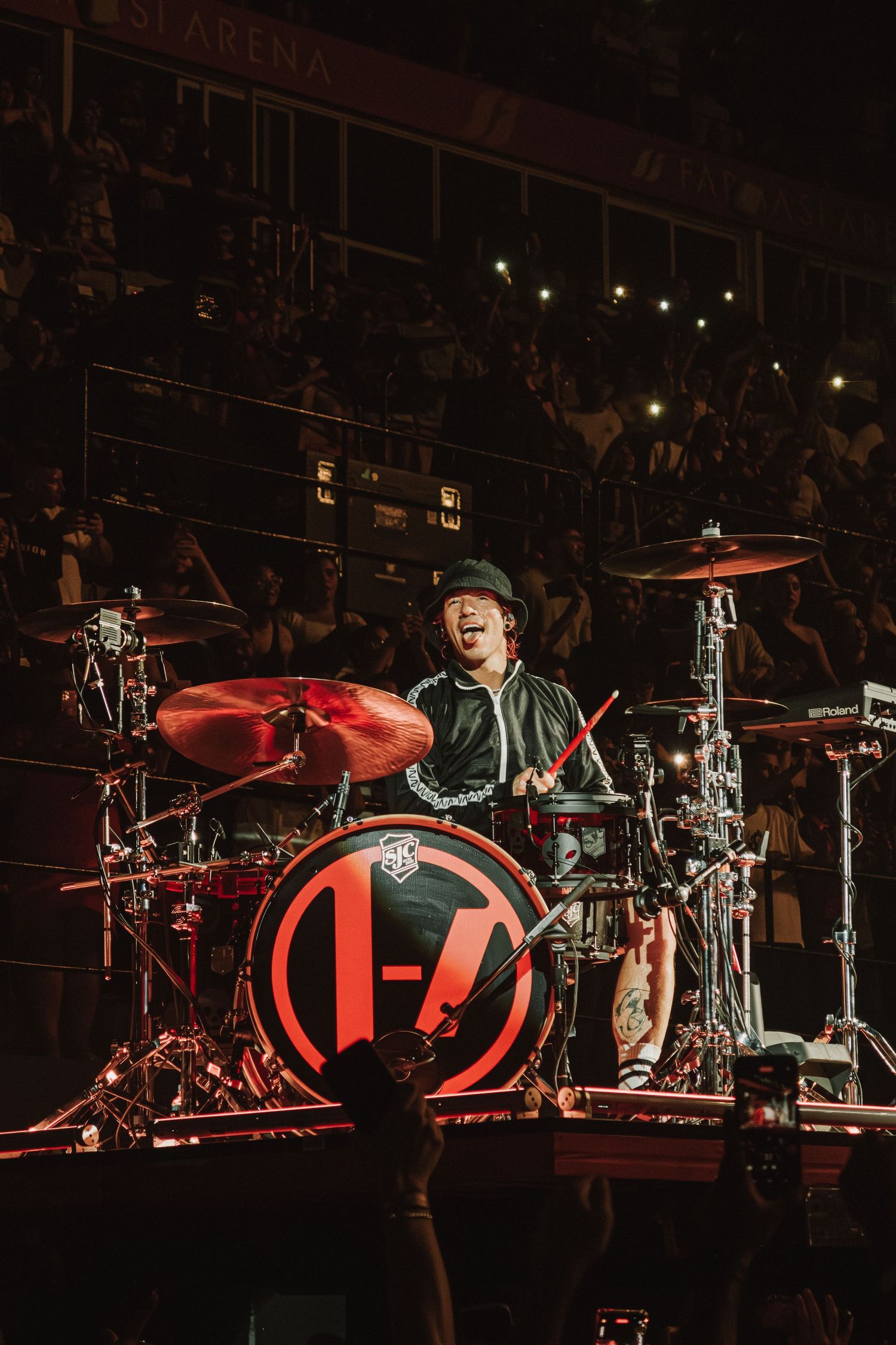Drummer performs energetically behind red-lit drum kit with band logo on arena stage.