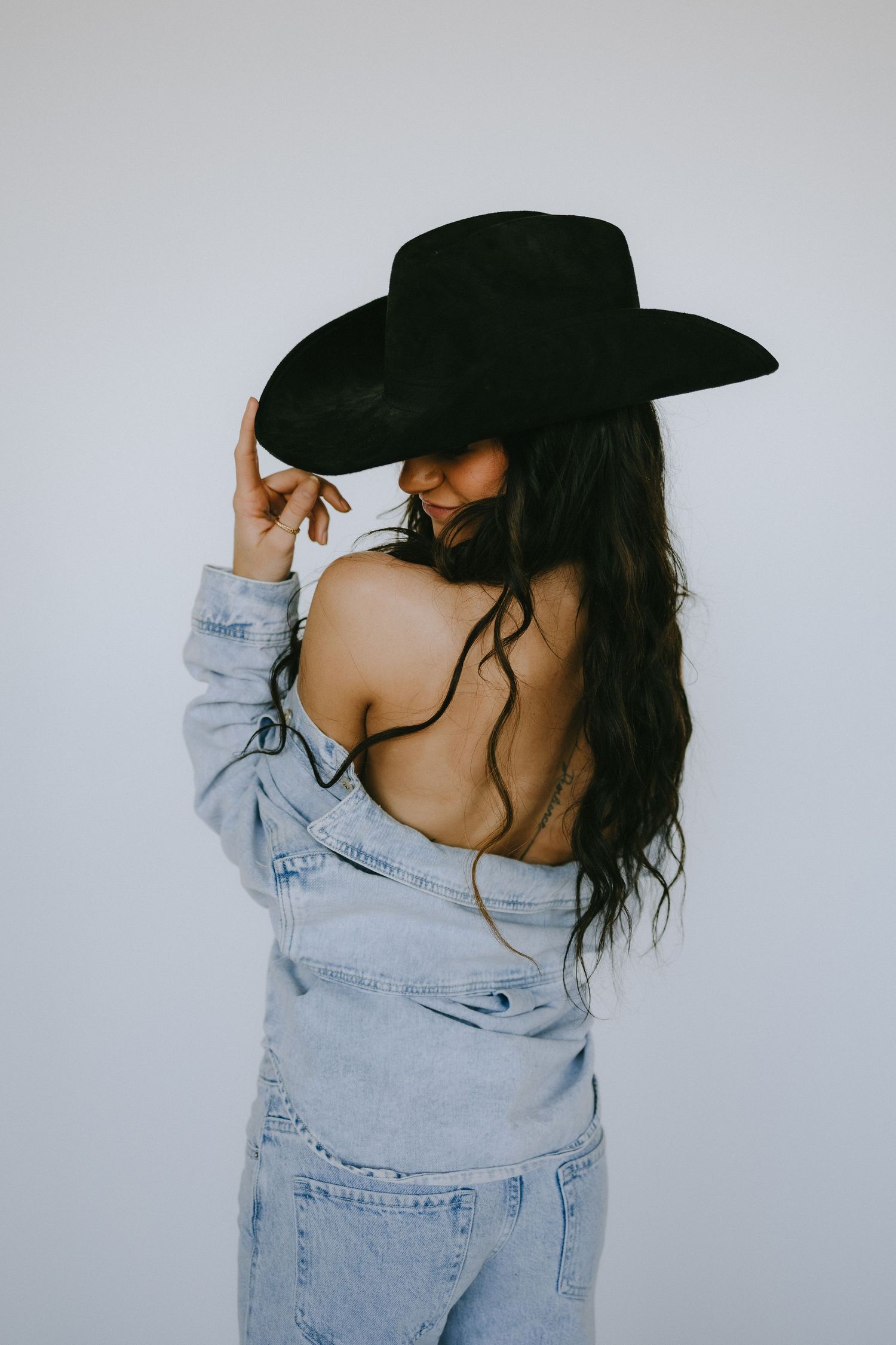 Artistic portrait of someone wearing denim and a black cowboy hat against white background.