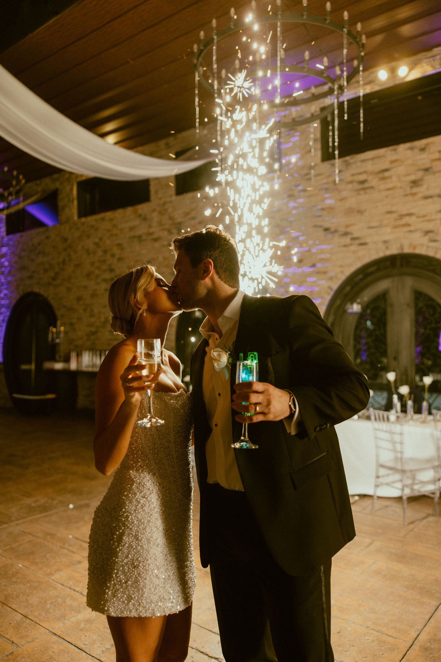 A romantic wedding reception moment with sparklers and champagne glasses under decorative lighting in a stone-walled venue.
