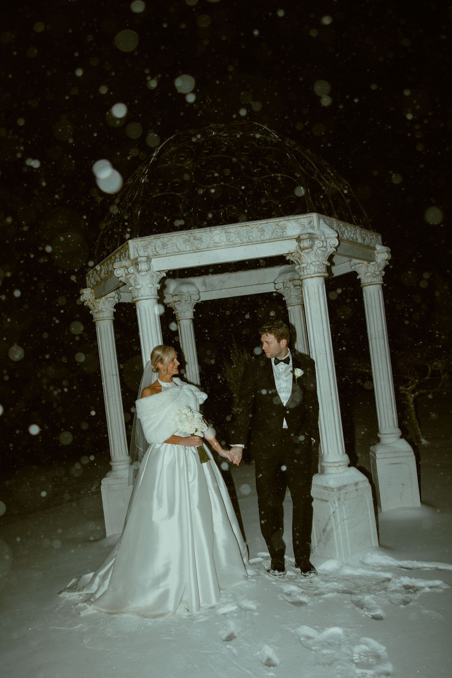A winter wedding scene with a couple posing by white columns in the snow.