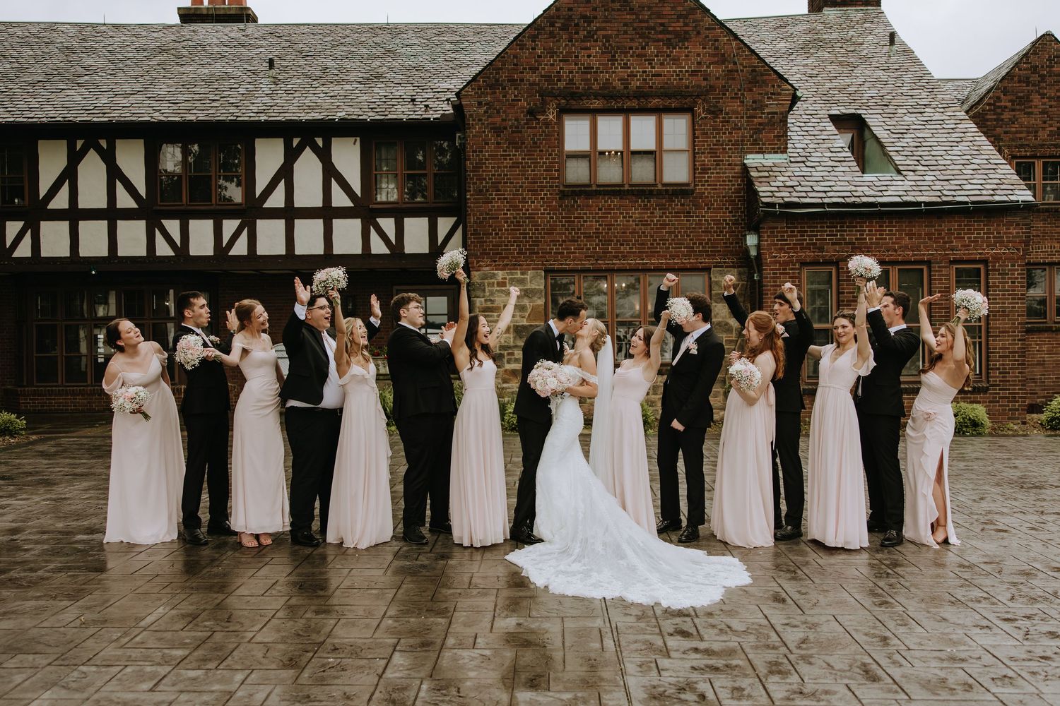 Wedding party in formal attire stands together outside a rustic brick and timber building on a rainy day.