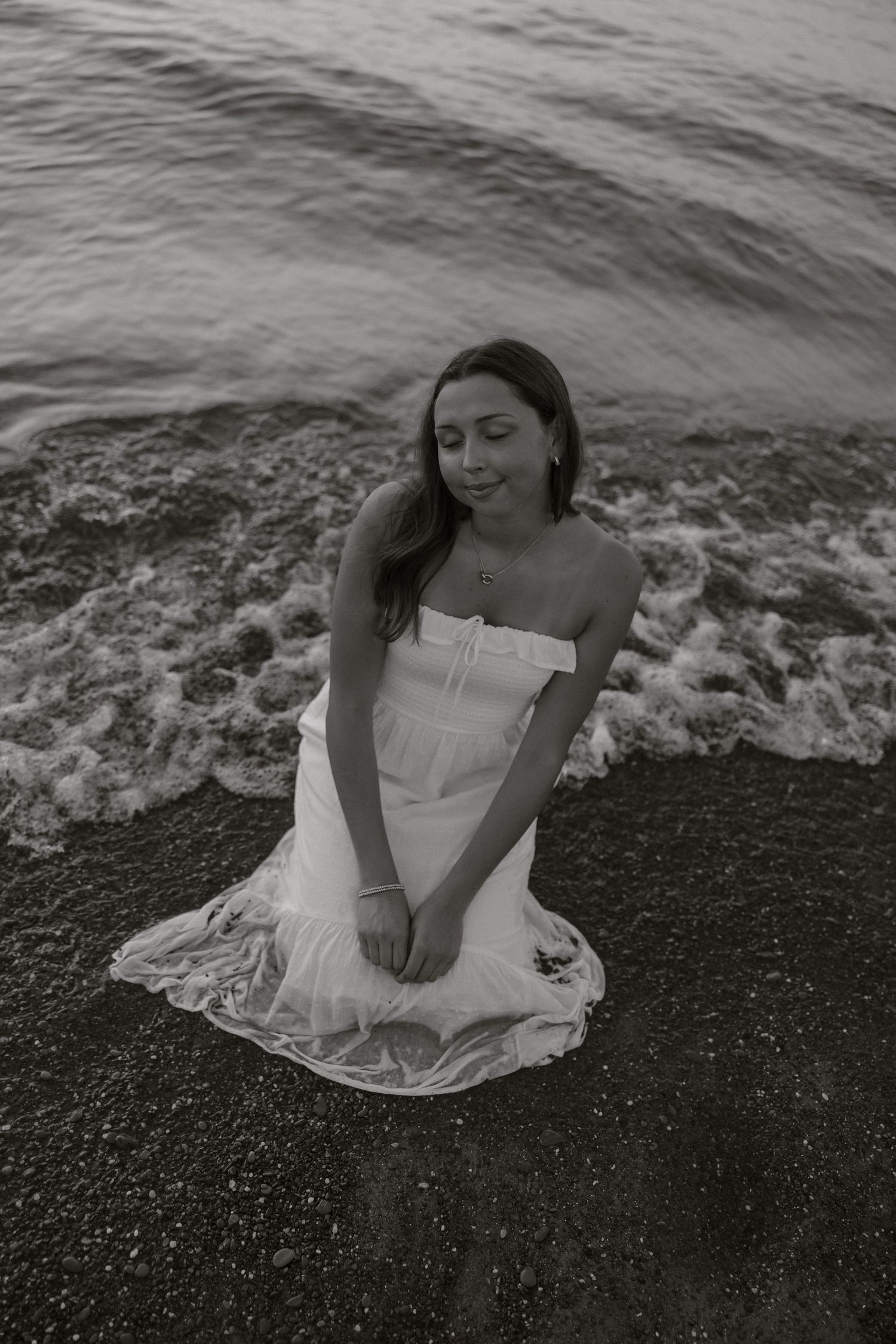 A woman in a white dress sits gracefully on a dark rocky beach as waves crash behind her in black and white.