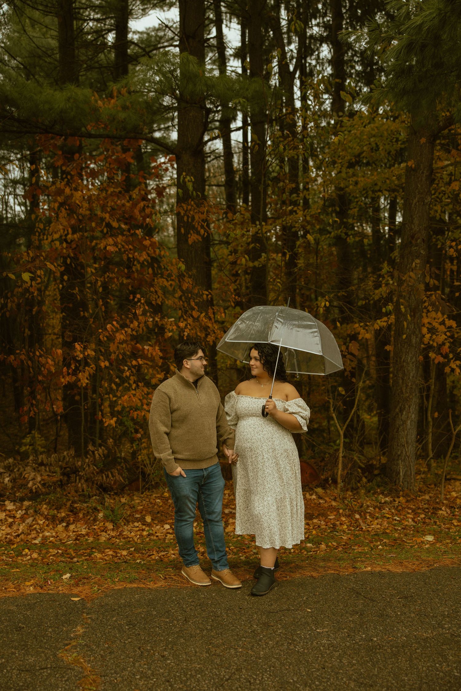 A couple sits on a curb under a clear umbrella surrounded by autumn foliage in a romantic fall photoshoot.