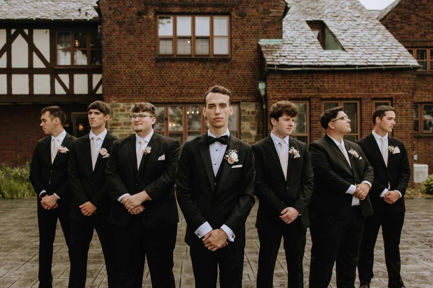 A row of groomsmen in black tuxedos stand together outside a brick building during a wedding.