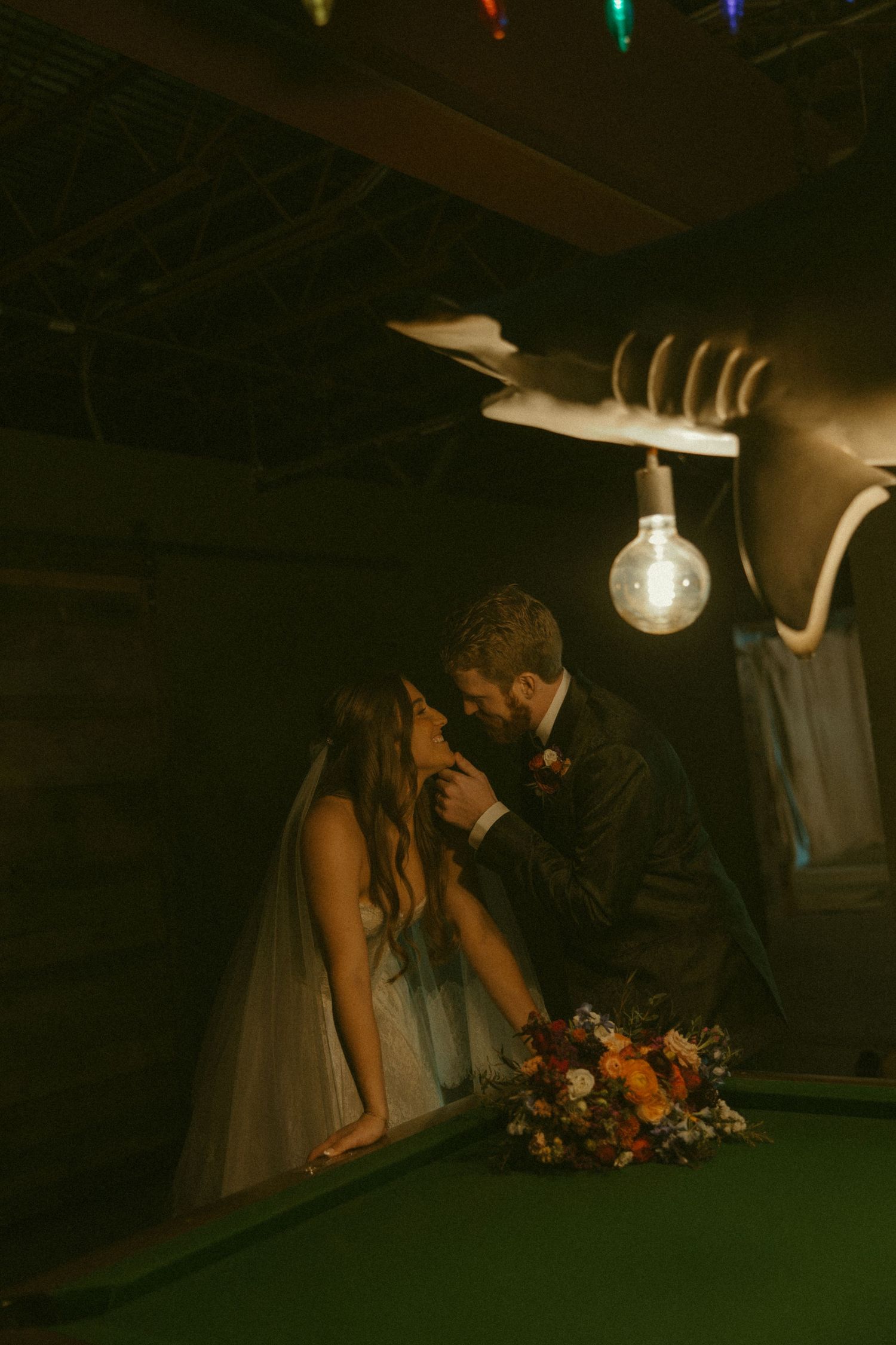 A romantic wedding couple shares an intimate moment in a dimly lit setting with colorful lights overhead.