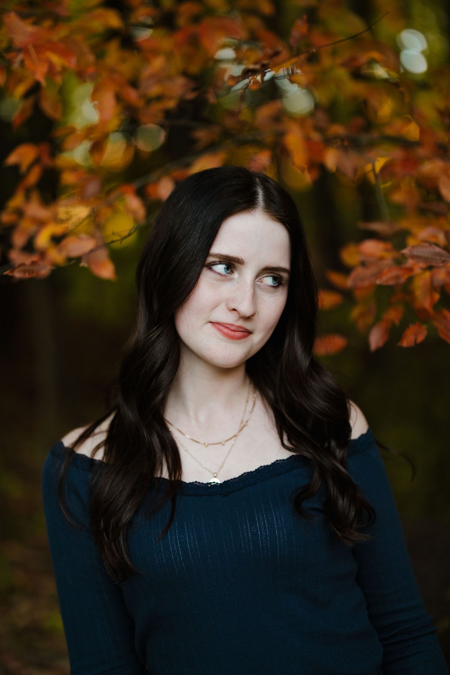 Portrait against autumn foliage wearing navy top and delicate necklace.