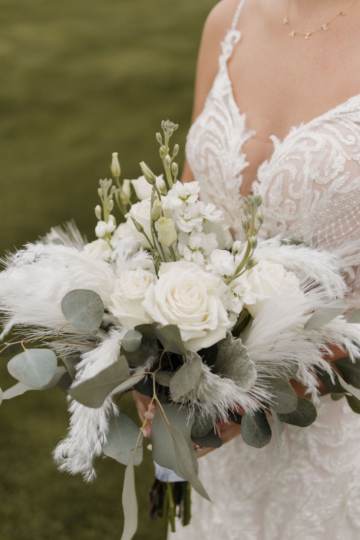 An elegant white wedding bouquet with roses, eucalyptus and feather accents against a lace dress.