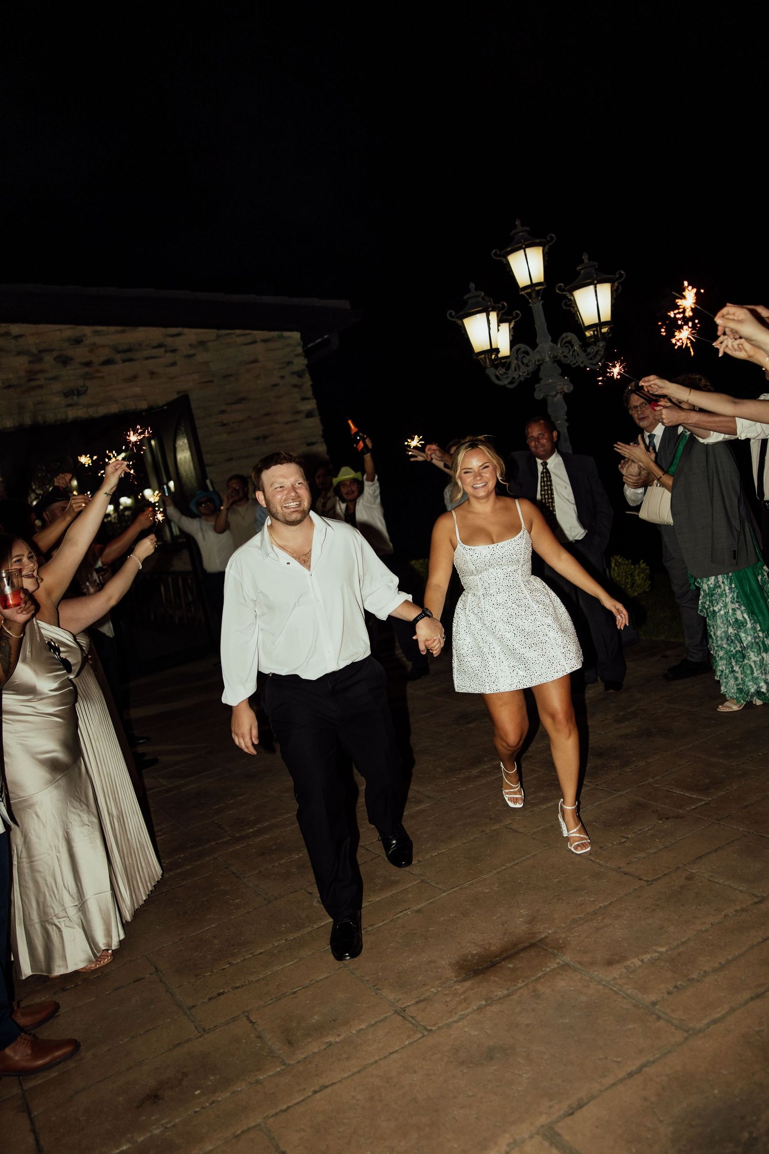 Wedding guests celebrate on a dance floor at night with illuminated lamp posts in the background.