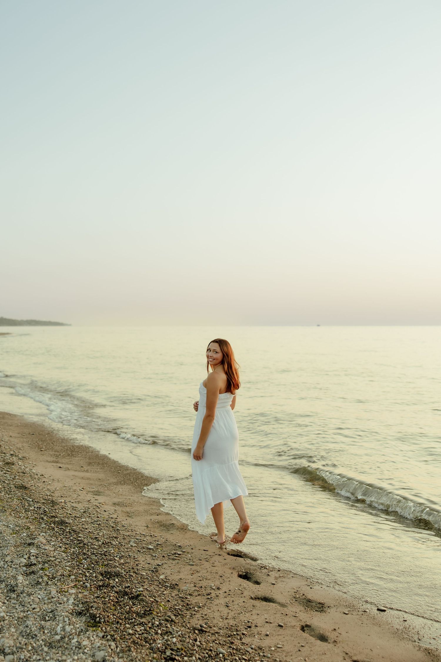 A person in a flowing white dress walks barefoot along a peaceful beach shoreline at sunset.