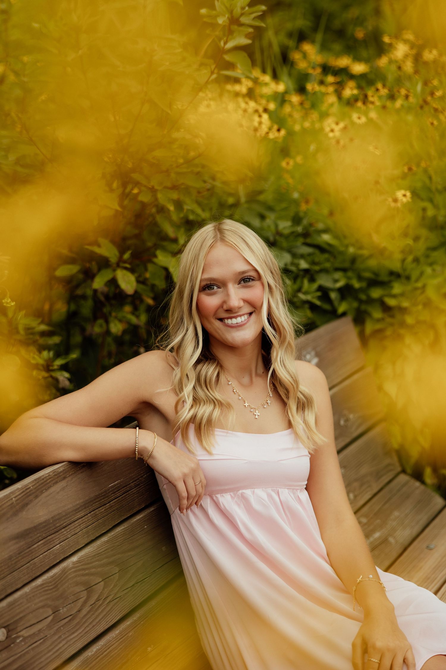 Summer portrait in a light pink dress sitting on wooden deck with golden sunlight and greenery in background.