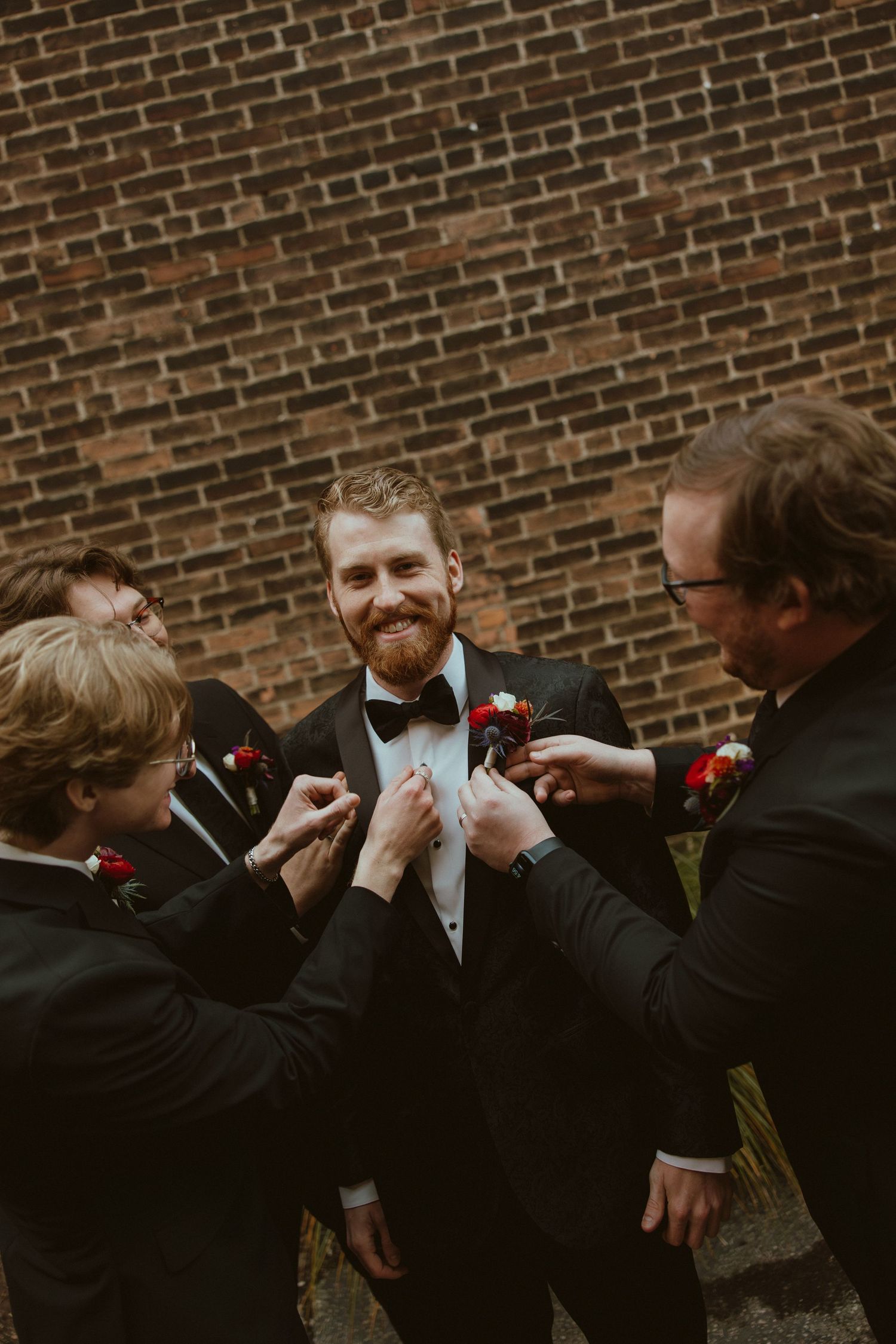 A sequence of groomsmen helping adjust a bow tie against a brick wall background.