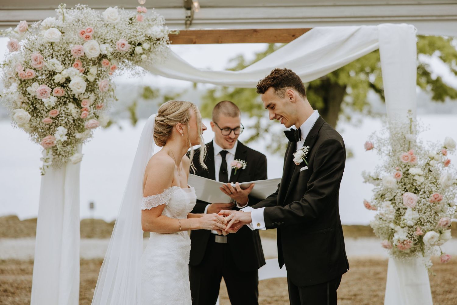A romantic outdoor wedding ceremony takes place under a white pergola decorated with pink and white floral arrangements.