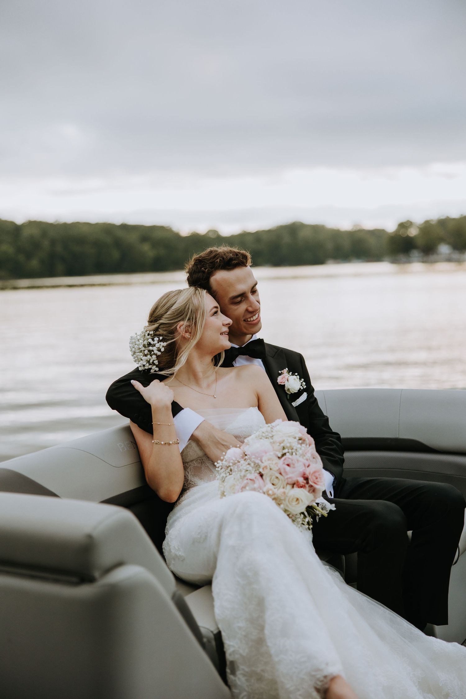 Newlywed couple sharing an intimate moment in a convertible car by a lake with a cloudy sky backdrop.