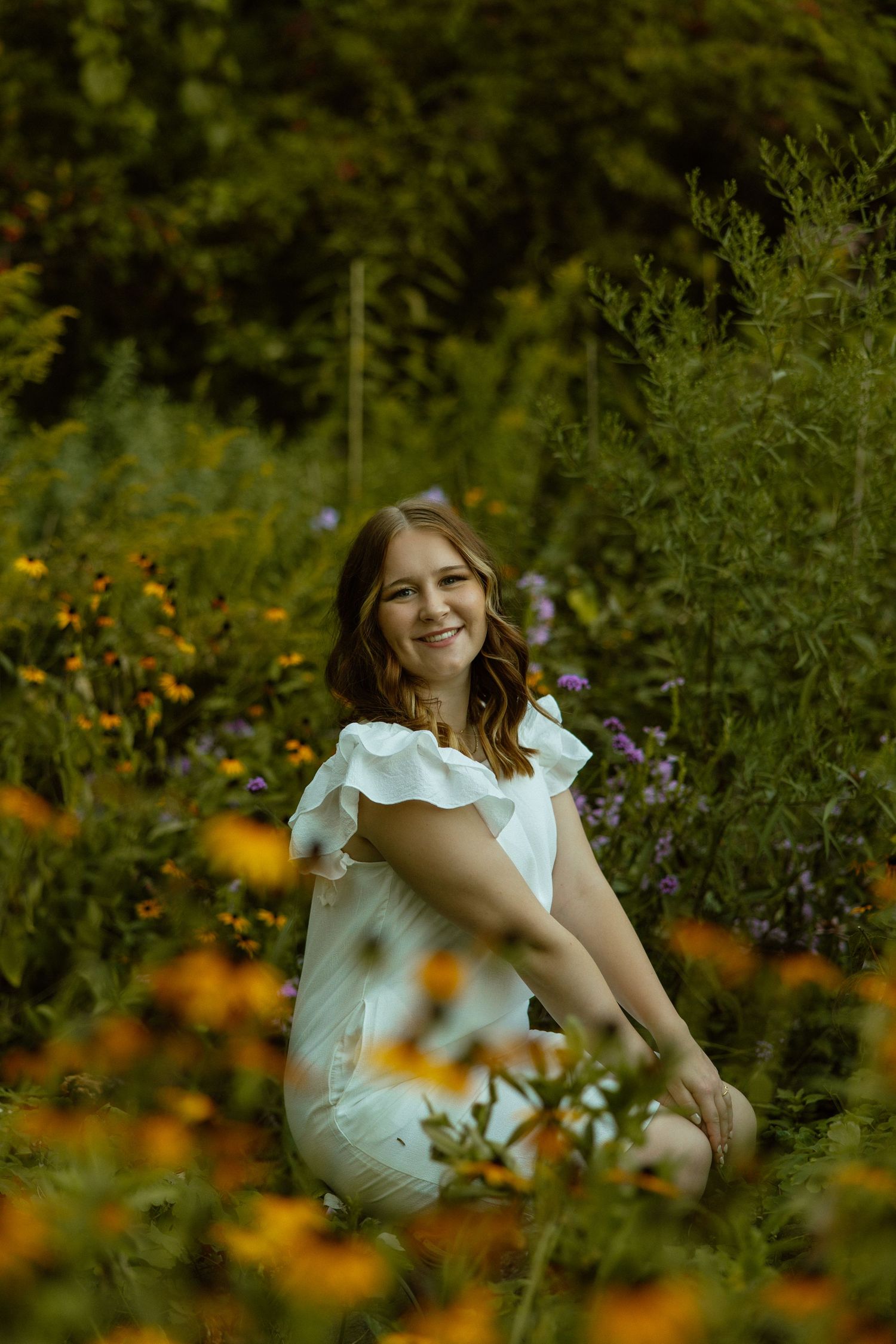 A person in a white dress sits among blooming wildflowers in a lush garden setting during golden hour.