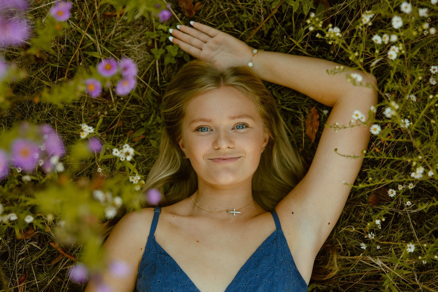 A person in a blue top poses in a garden setting with purple flowers and natural sunlight.