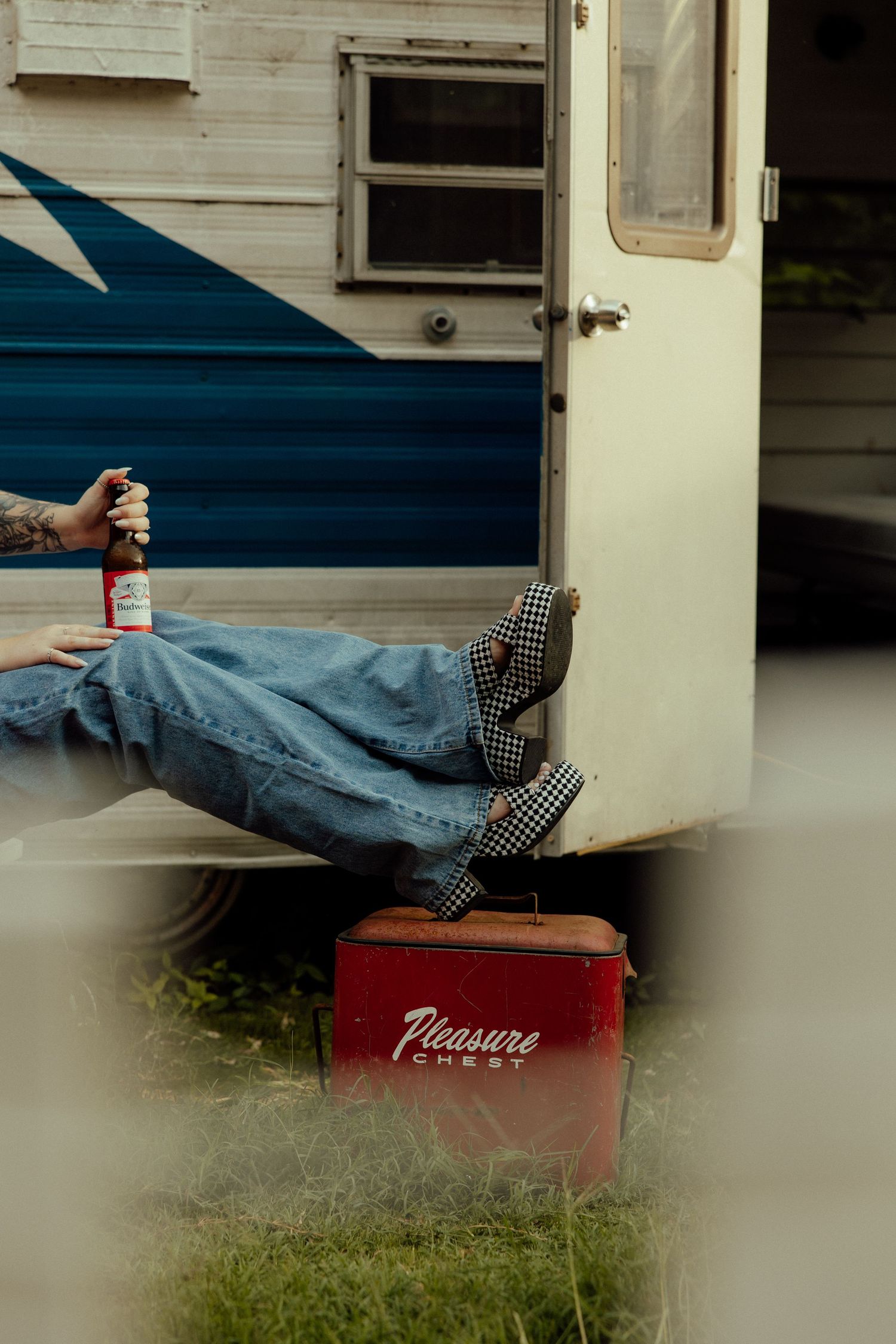 A red vintage cooler sits in front of a white trailer on grass with legs resting on top.