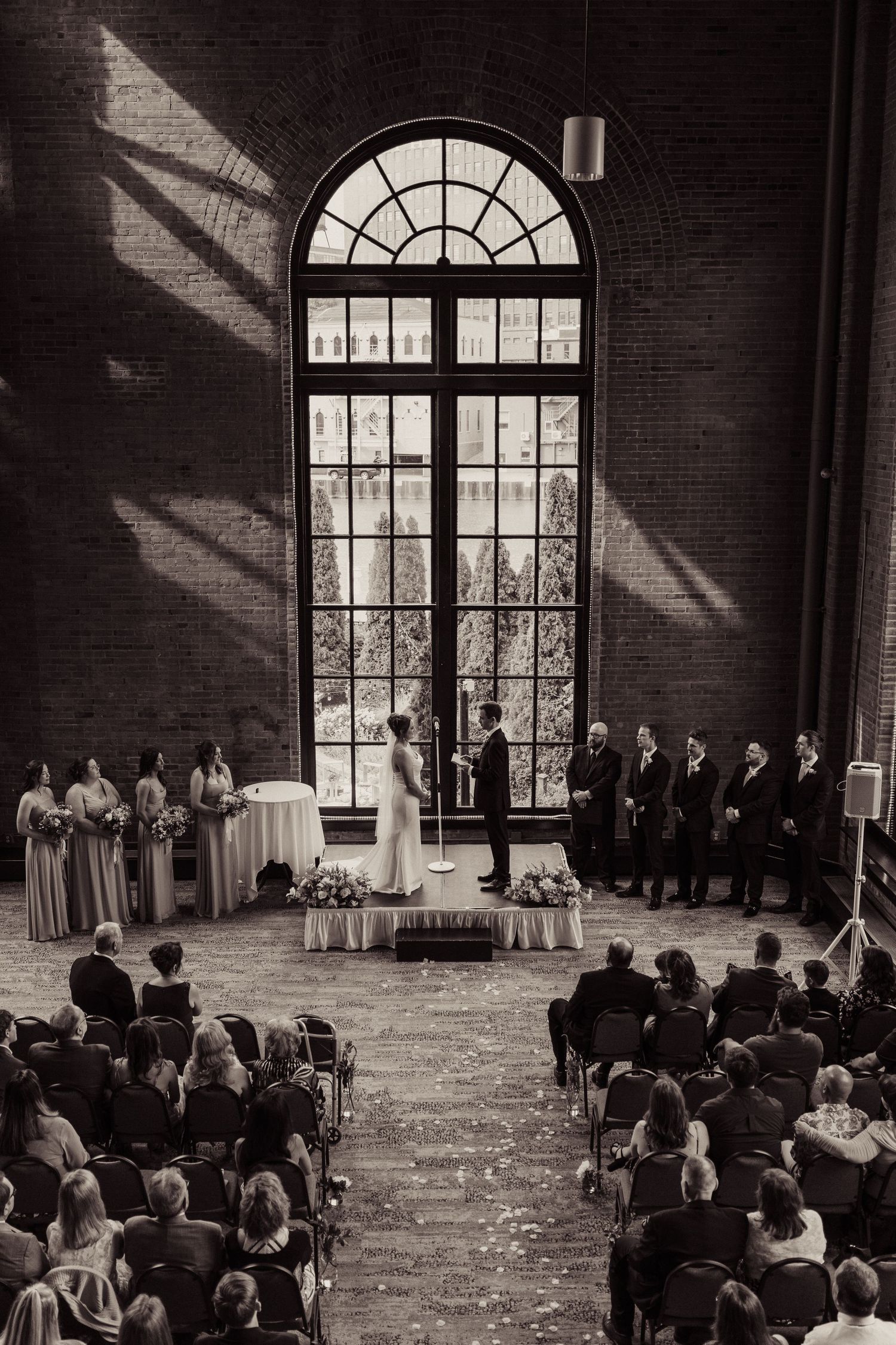 A black and white photo of an indoor wedding ceremony with a dramatic arched window and gathered guests.