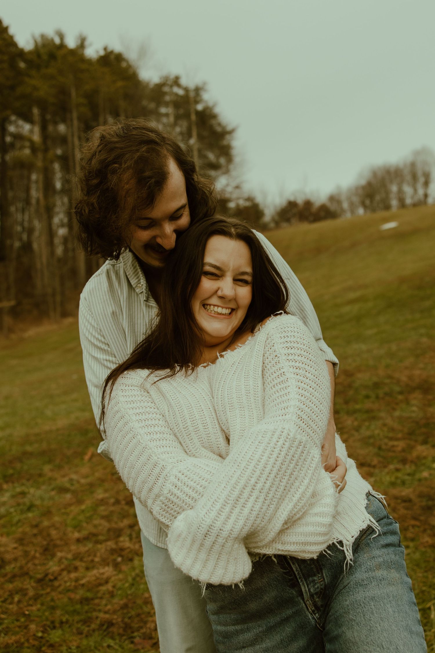 A couple embraces and laughs together on a grassy hillside with pine trees in the background during golden hour.