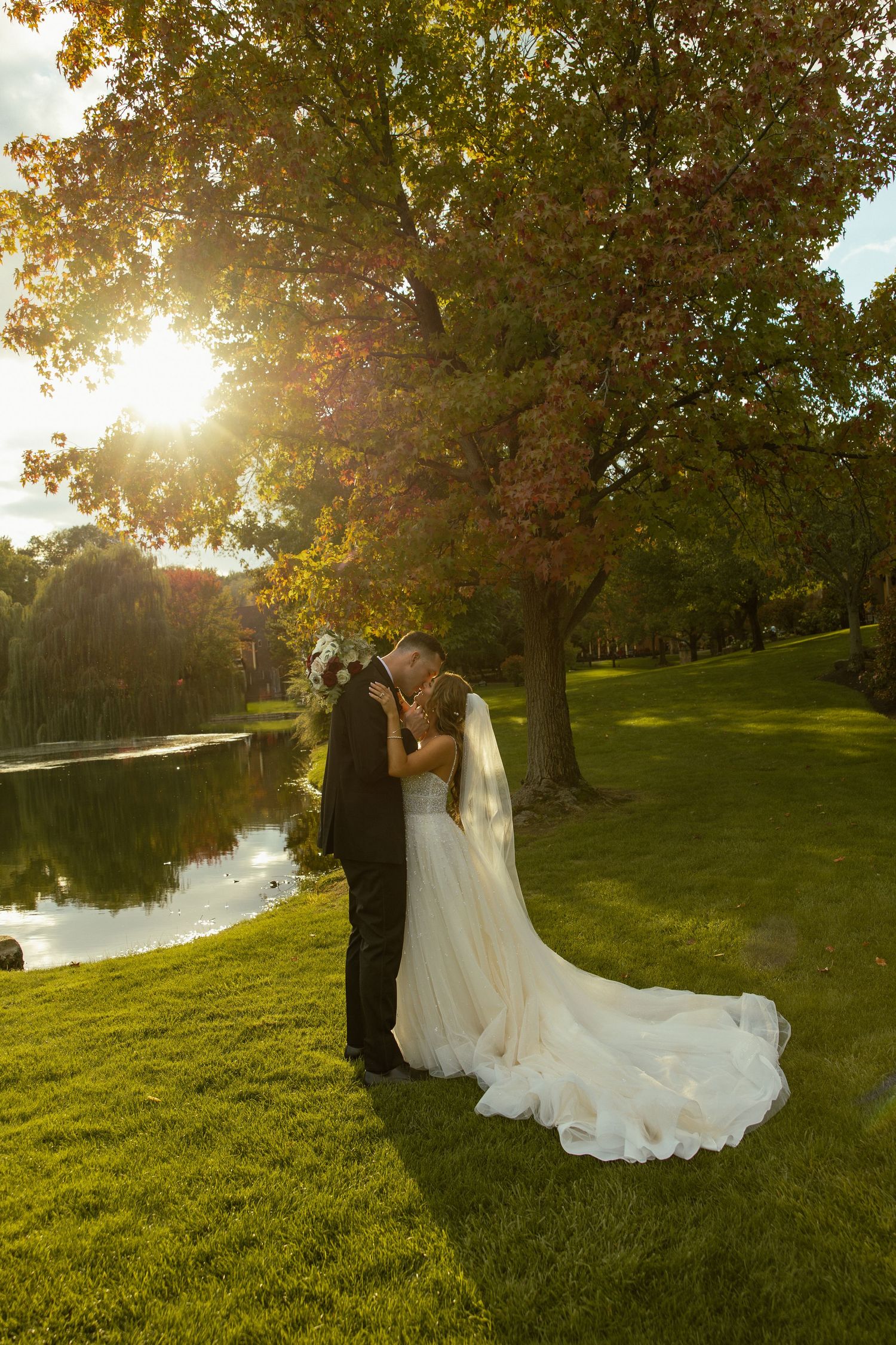 A romantic sunset wedding photoshoot by a lake with golden sunlight streaming through trees onto a grassy lawn.