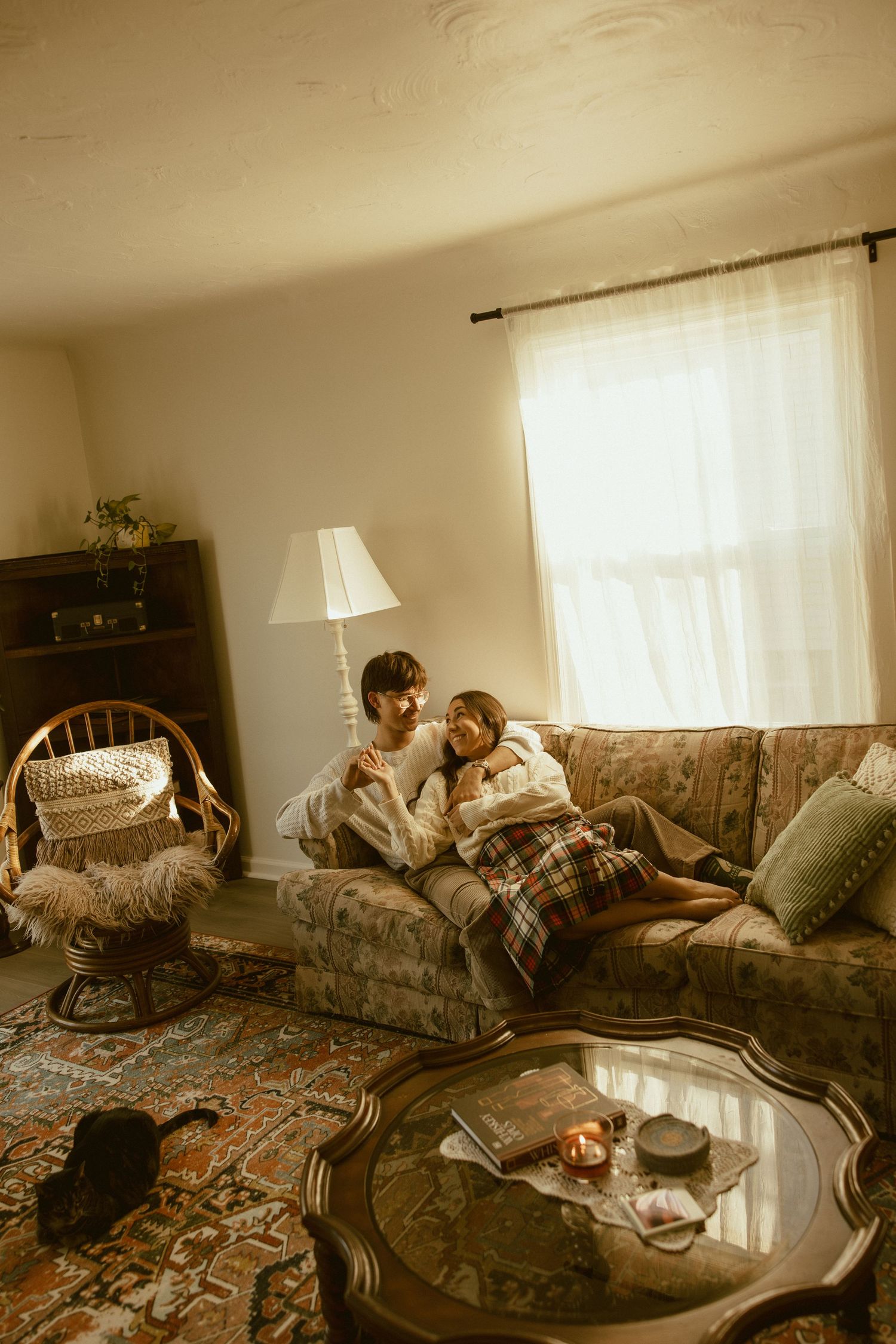 A cozy vintage living room with a floral couch, wicker chair, ornate coffee table and soft window light creating a warm atmosphere.