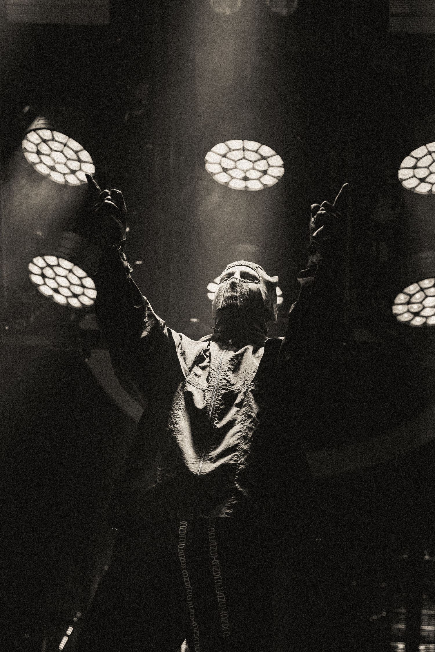 Black and white concert shot of performer with raised arms under spotlight array.