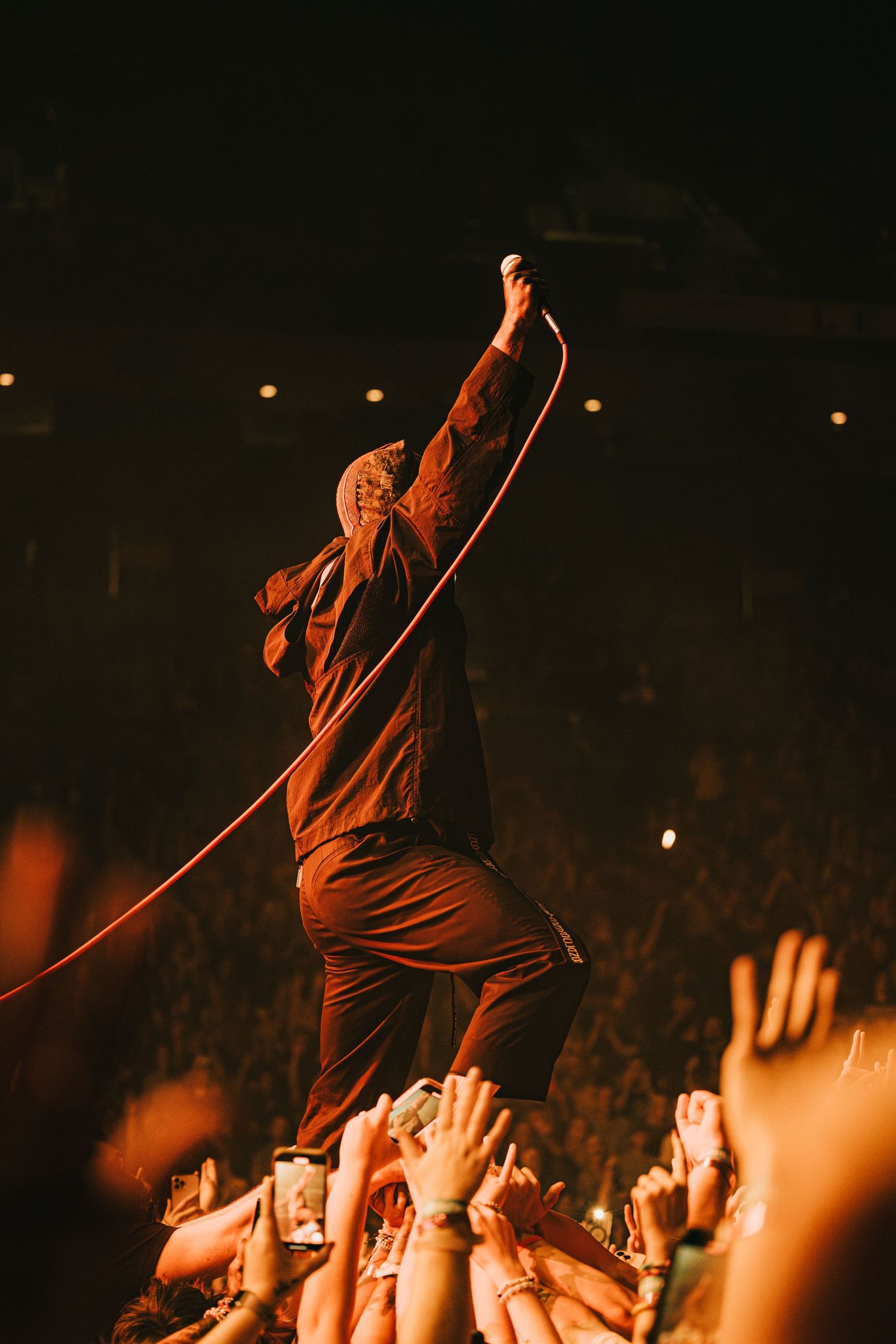 Performer in red lighting reaching up while crowd raises hands during concert.