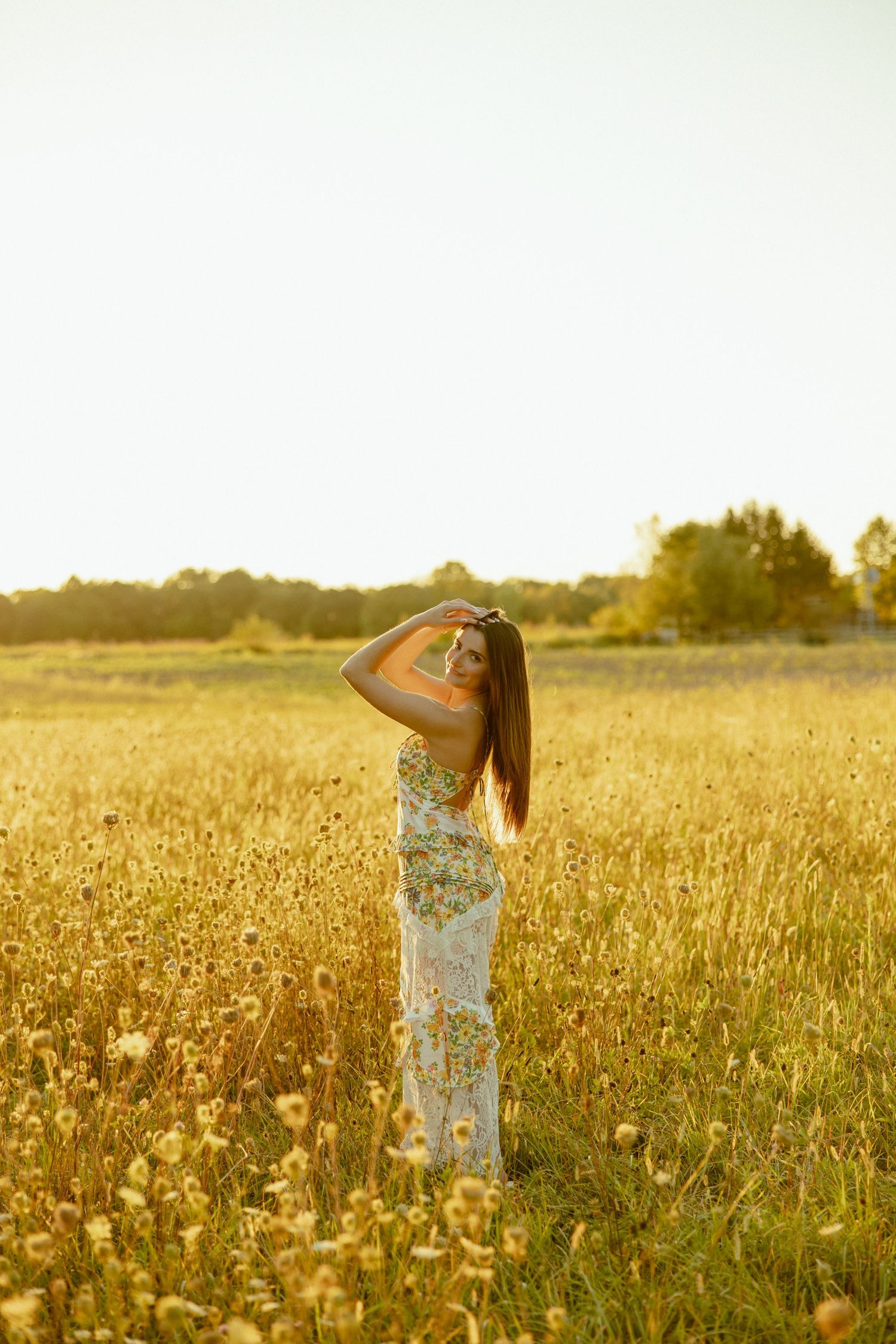 Person in floral dress stands in golden wheat field at sunset stretching arms upward in joyful pose.