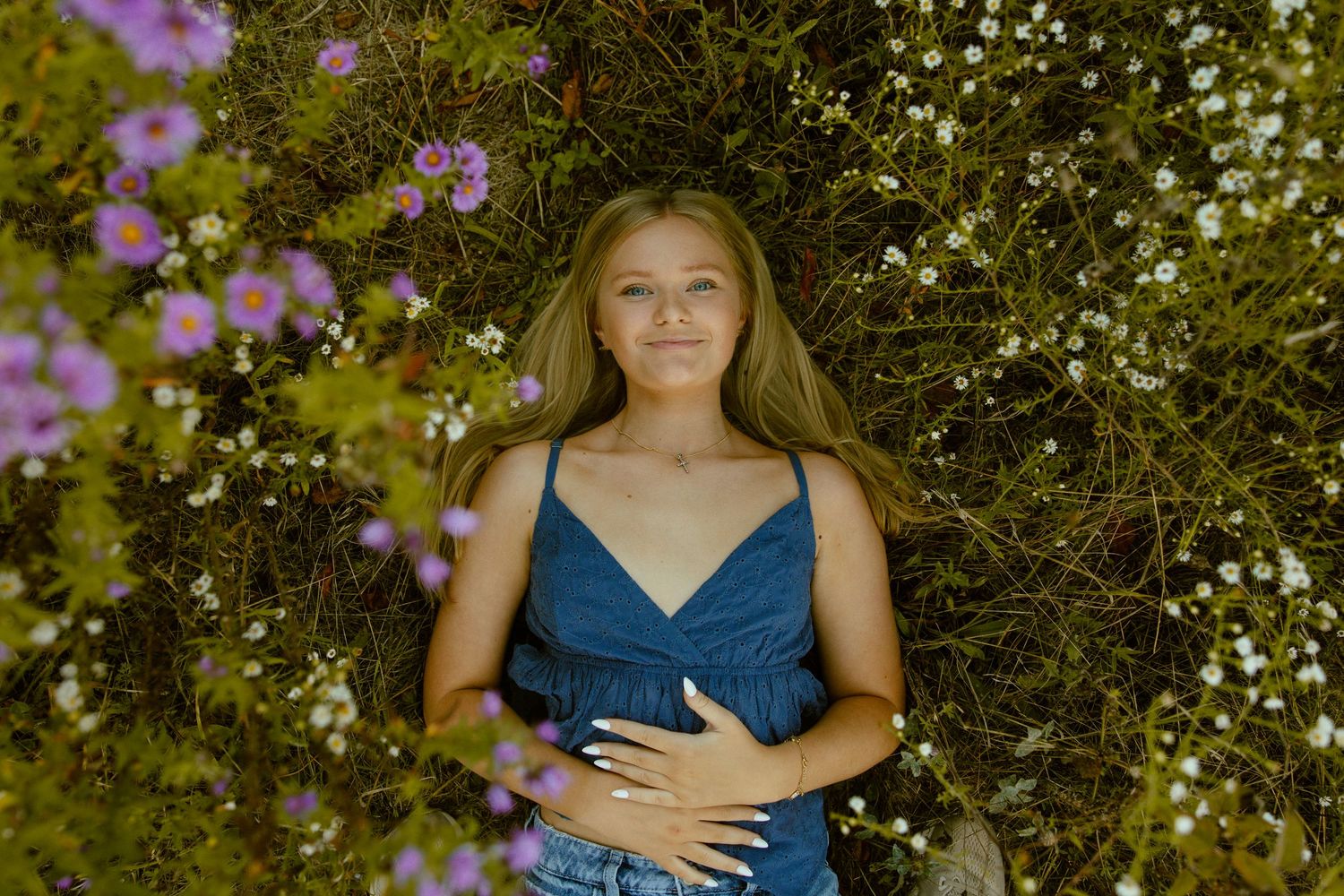 A person in a blue top poses among blooming flowers and greenery in a garden setting.