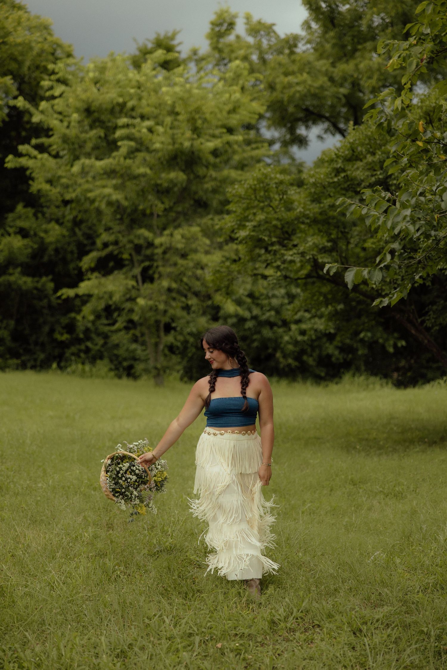Figure in a white fringe dress twirls playfully in a sunlit meadow surrounded by trees.