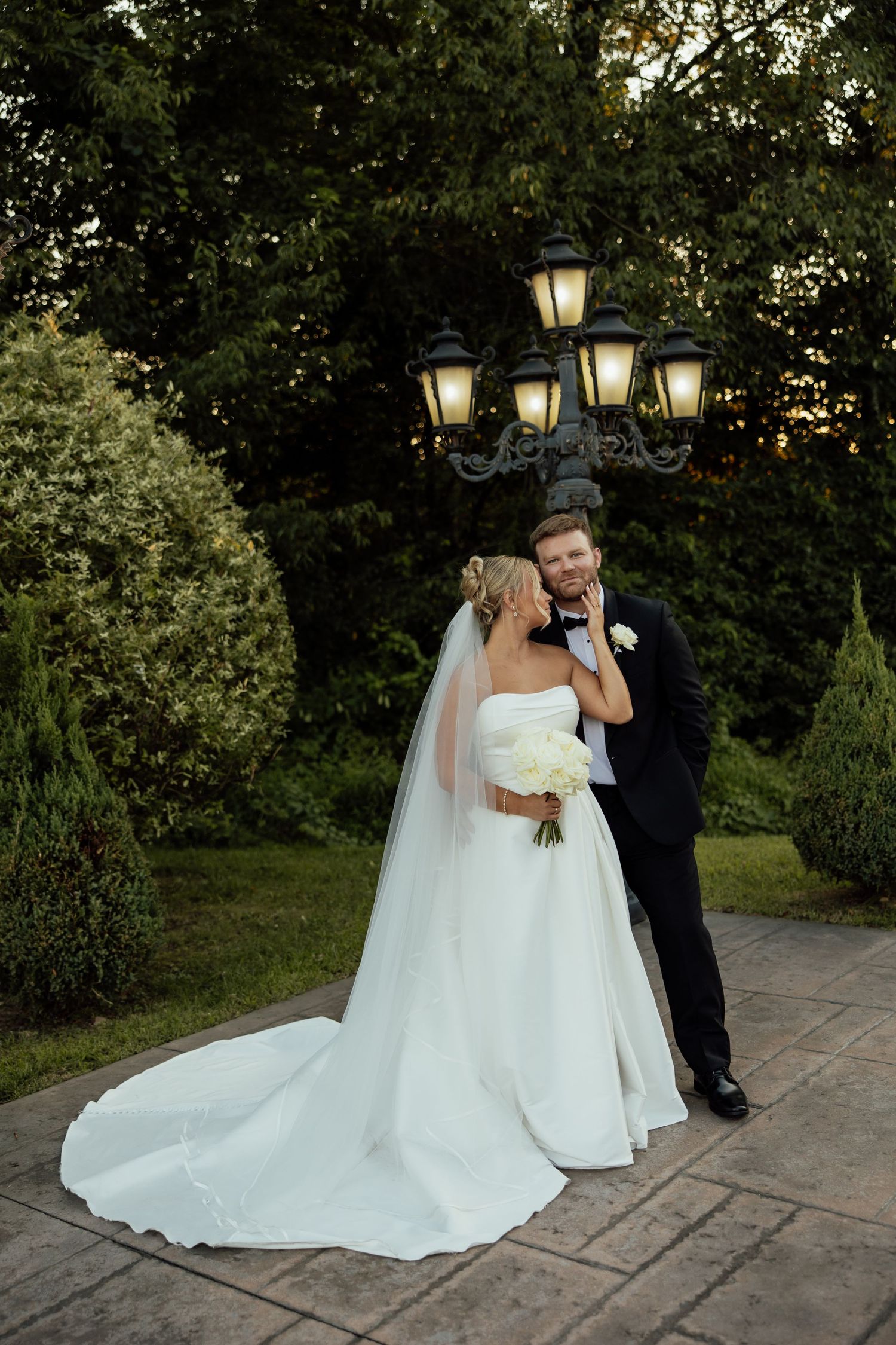 A romantic wedding couple poses under an ornate lamp post in a garden setting at dusk.