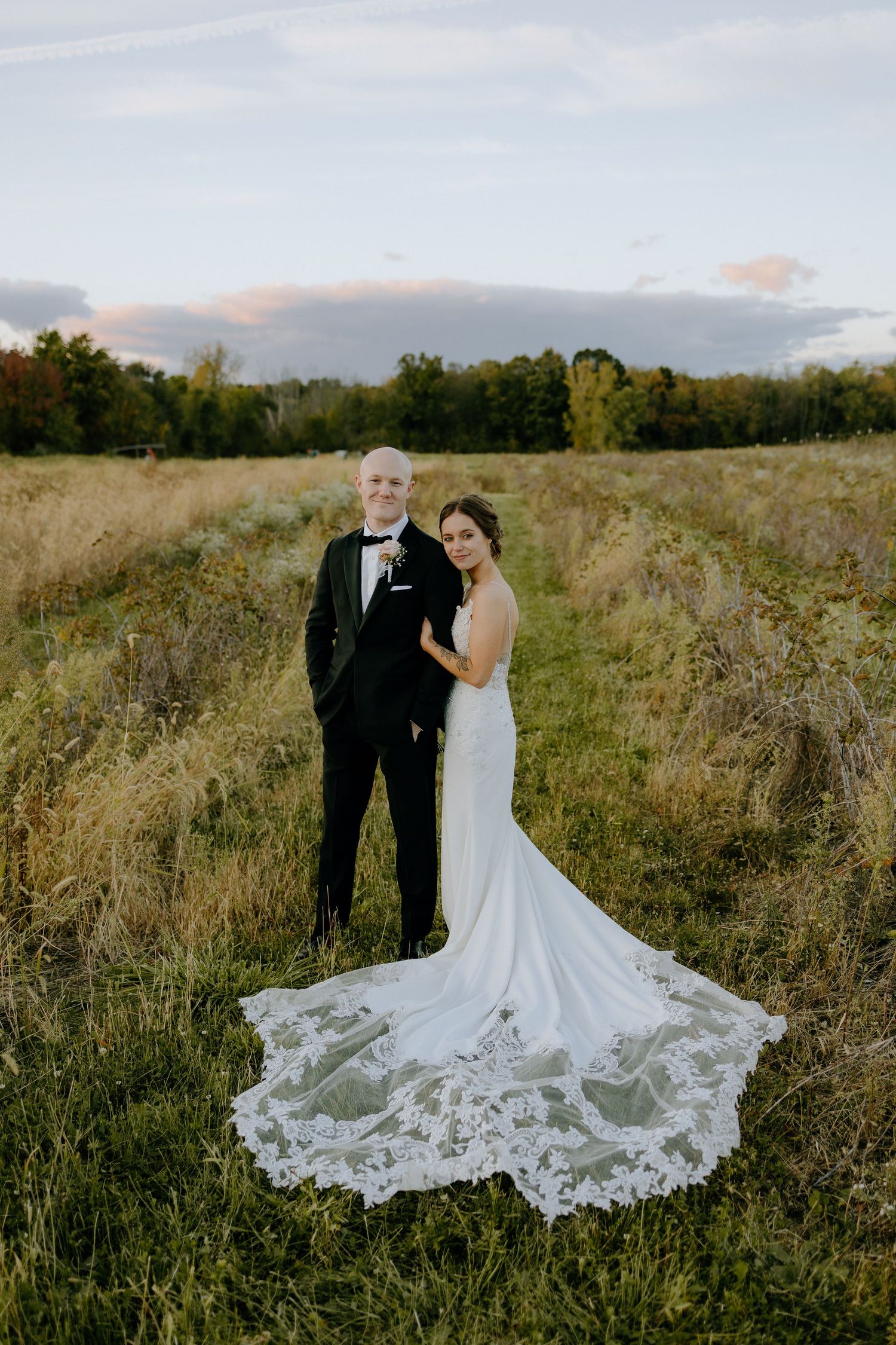 A newlywed couple stands in a golden field at sunset, with the bride wearing a flowing lace dress with long train.