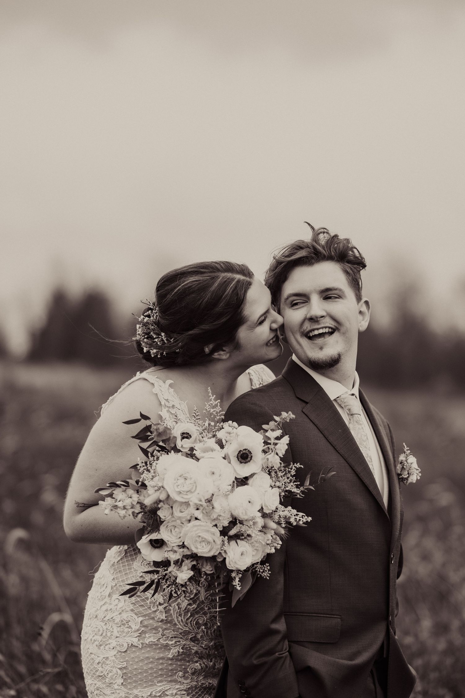 A black and white wedding photo showing a joyful moment in a field with a beautiful floral bouquet in focus.