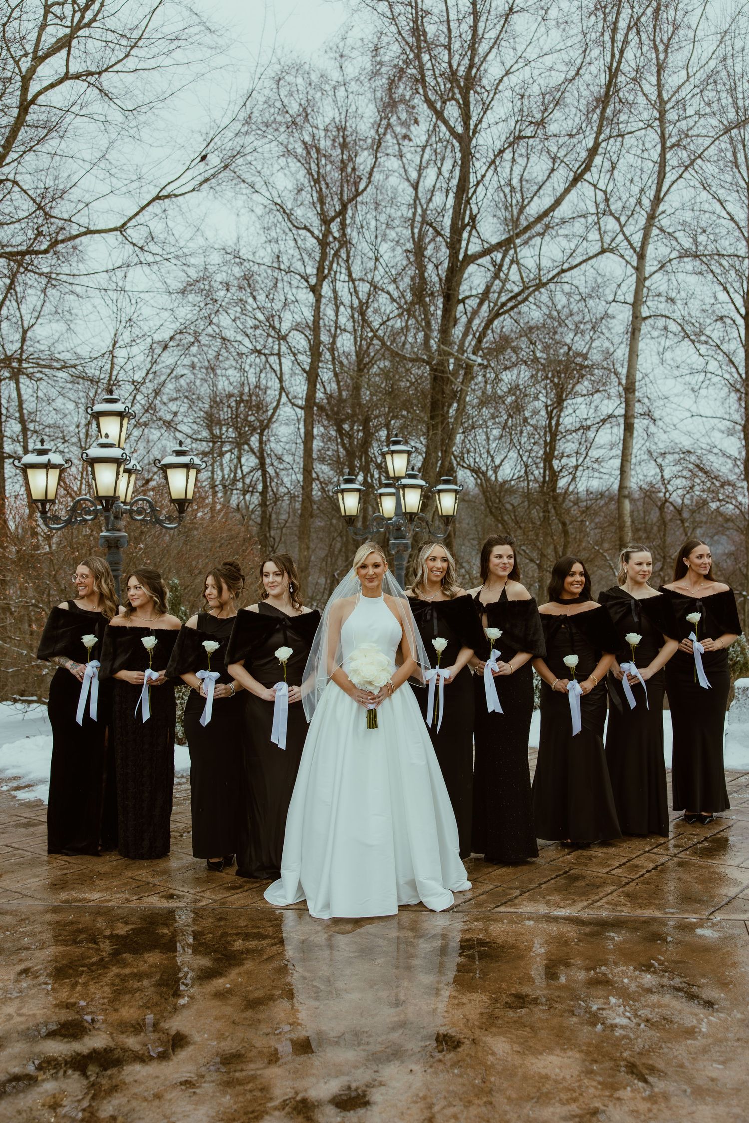 Wedding party in black dresses stands in a snowy outdoor setting with lanterns and bare trees.