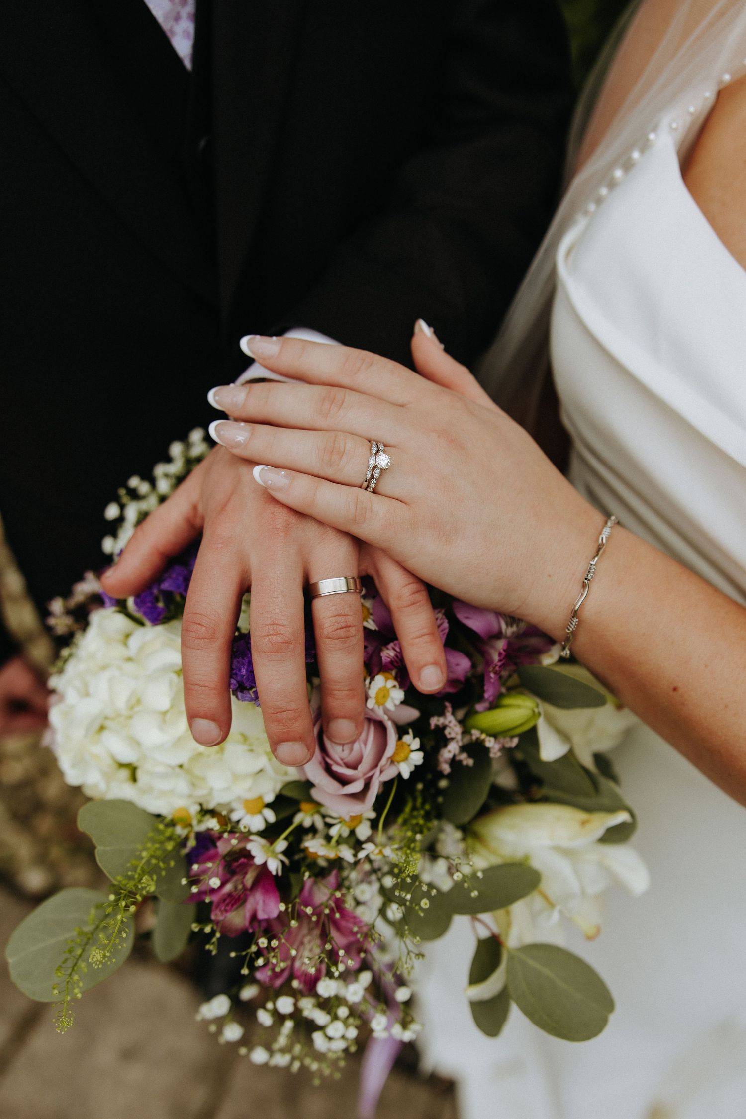 Close-up details of wedding rings and floral bouquet with white and purple flowers.