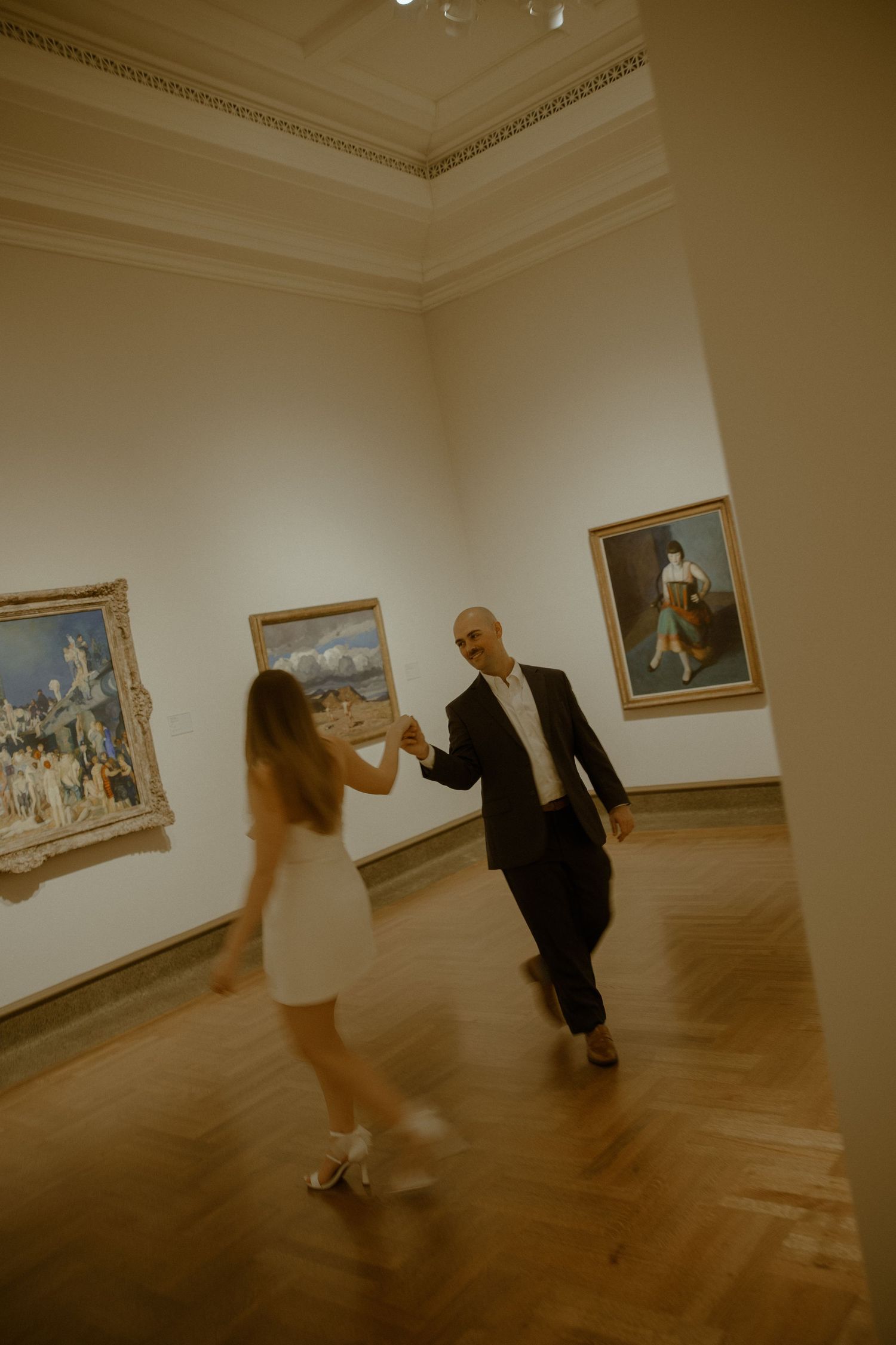 A couple dances together in an art gallery with classic paintings on display and hardwood floors.